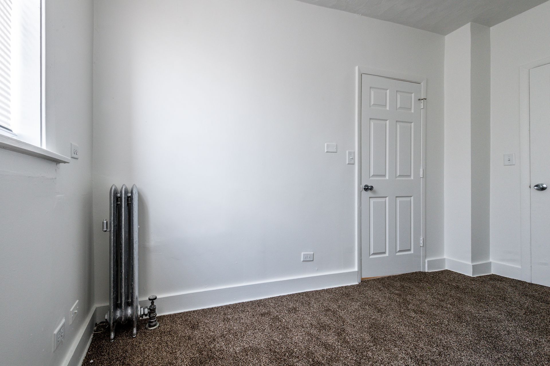 Empty room with white walls, a closed door, radiator, and carpeted floor.