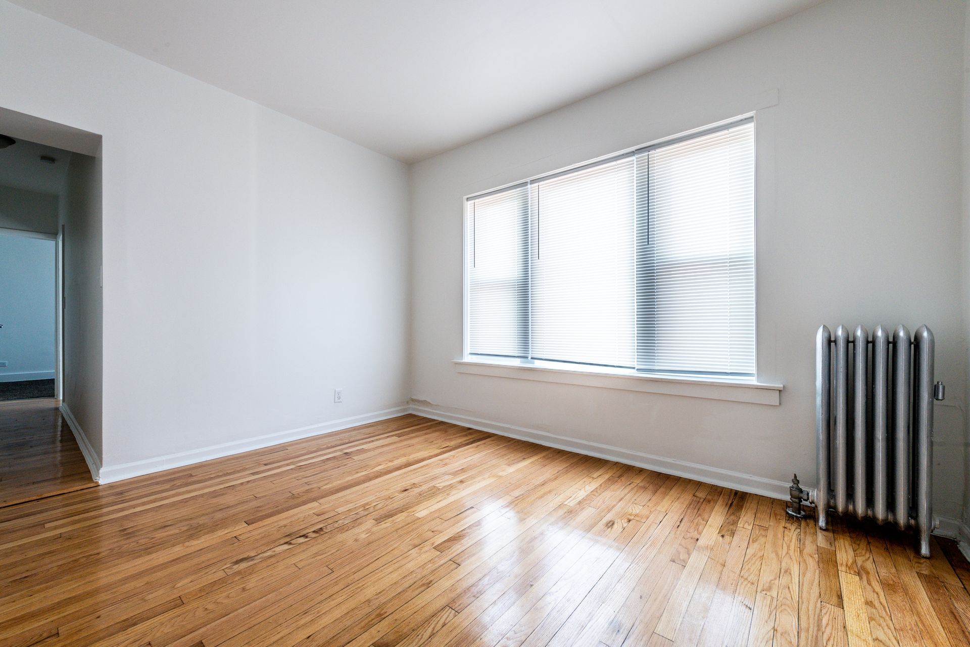 Empty room with wood floors, white walls, window with blinds, and radiator.