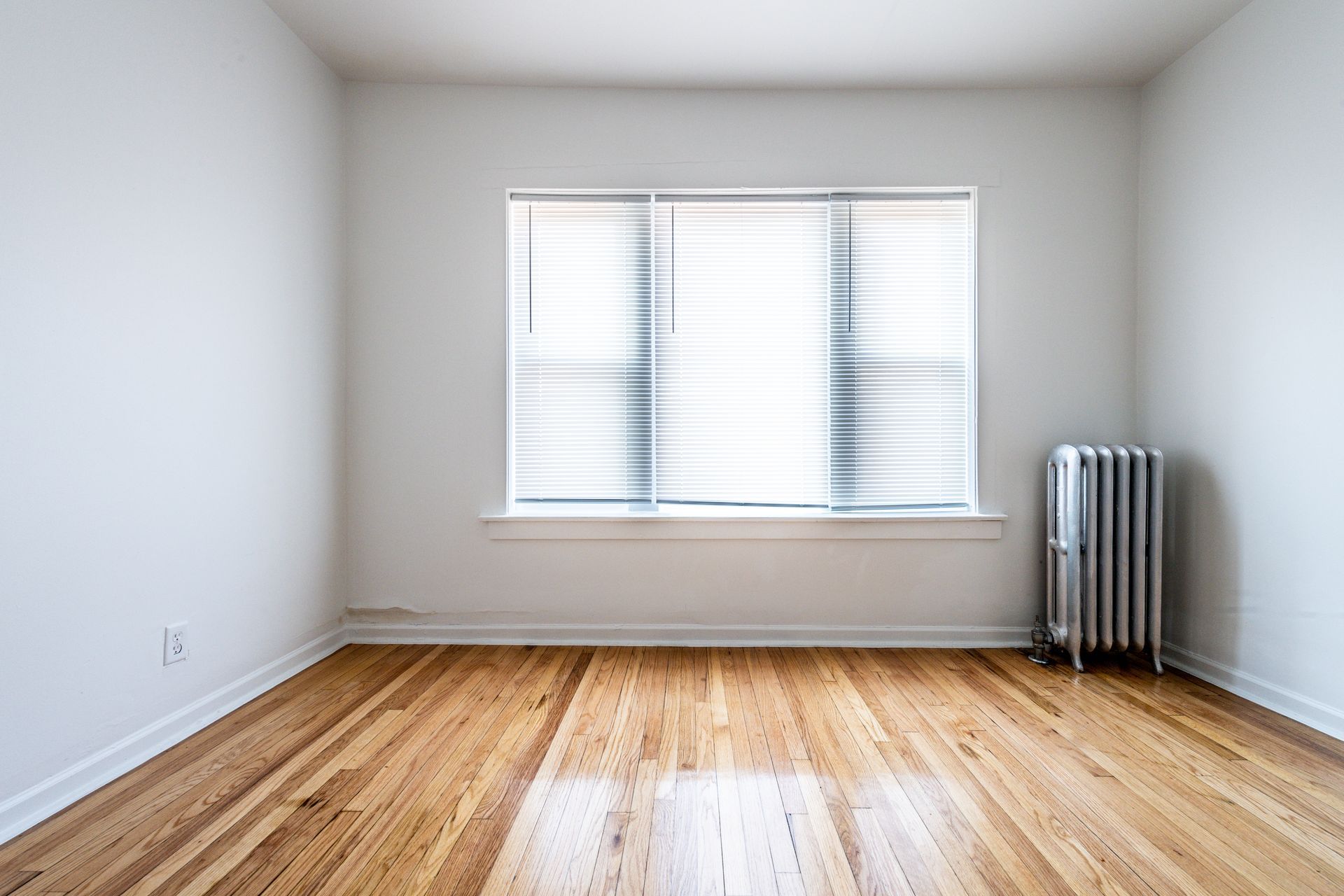 Empty room with wood floor, white walls, window with blinds, and radiator.