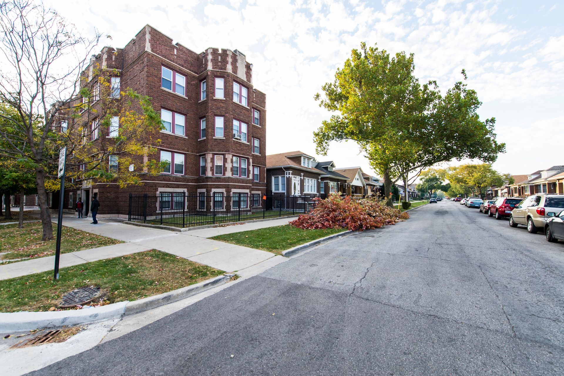 Brick apartment building on a residential street with cars parked along the side, and trees on the lawn.