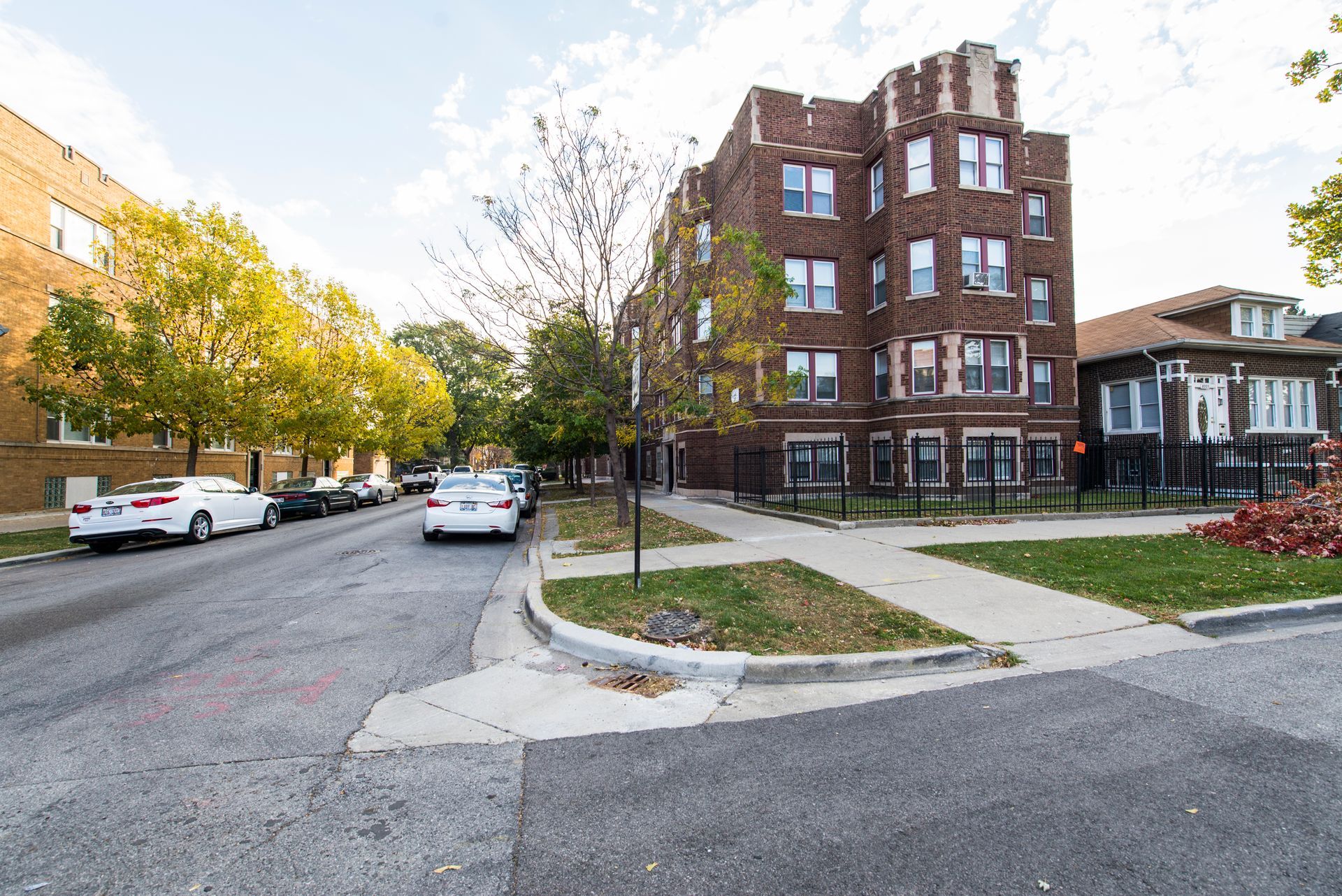 Street view of brick apartment building on corner lot, with parked cars and trees.