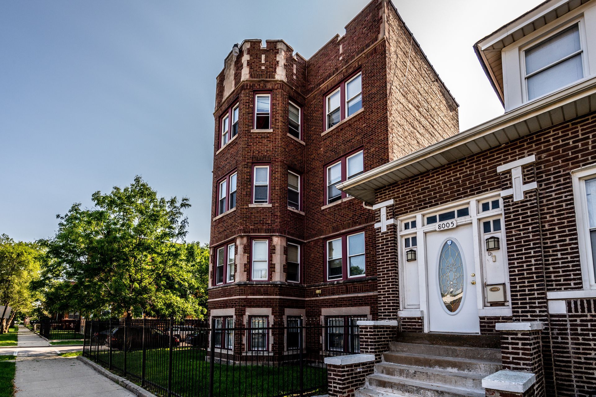 Brick apartment building with a turret, and a connected brick home, on a sunny day.