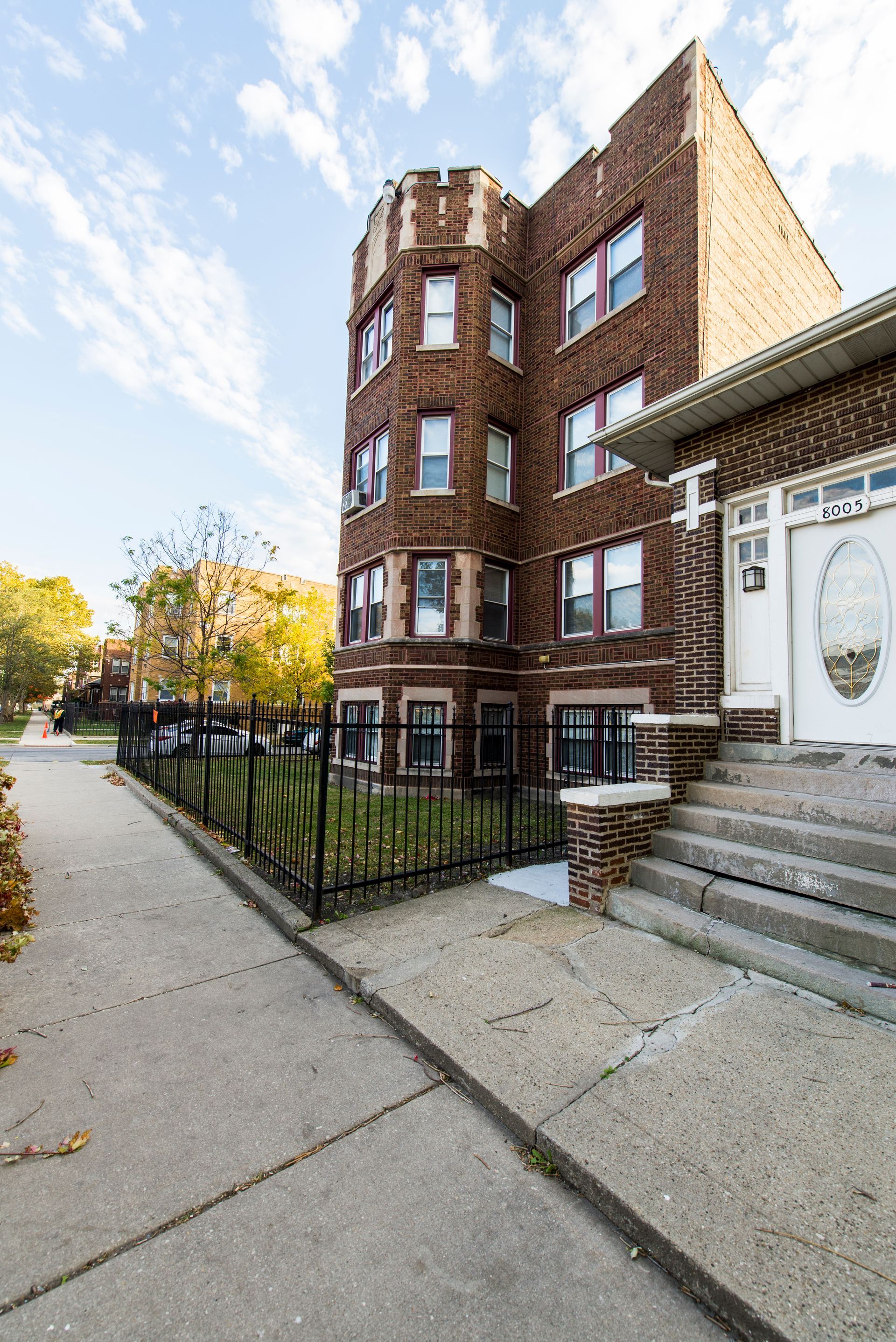 Brick apartment building with turret and wrought-iron fence on a sidewalk under a blue sky.