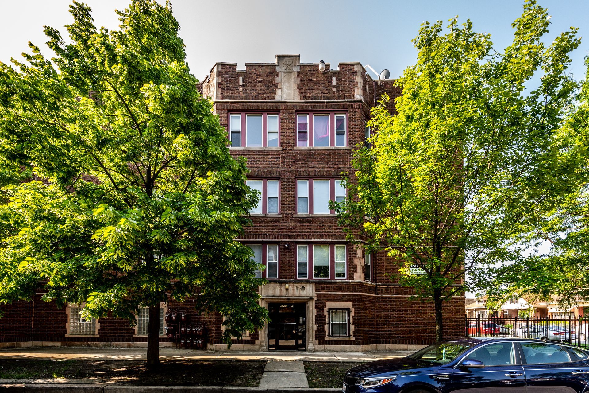 Brick apartment building with trees in front; blue car parked in the street.