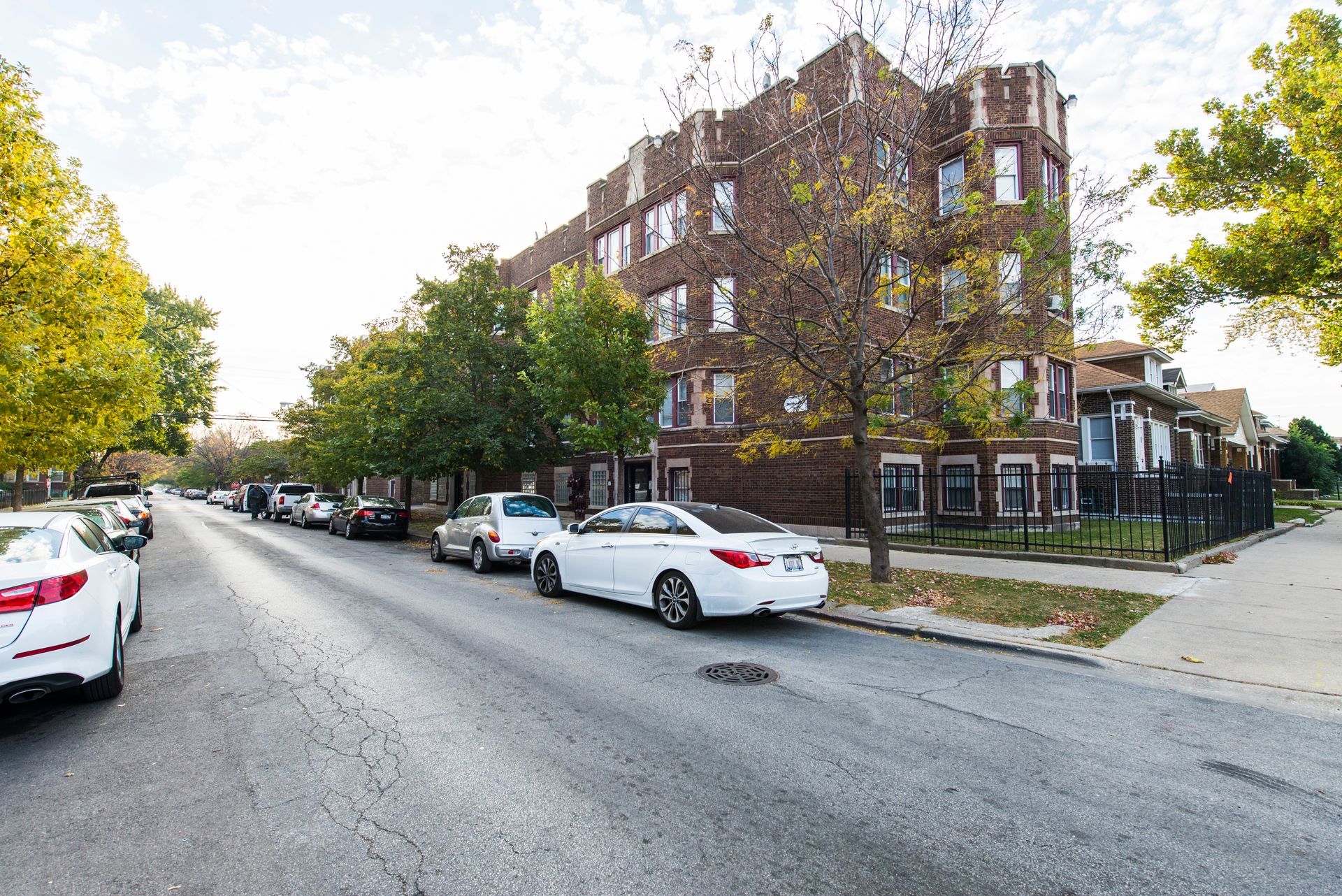 Street view of a brick apartment building with parked cars, and trees on a sunny day.
