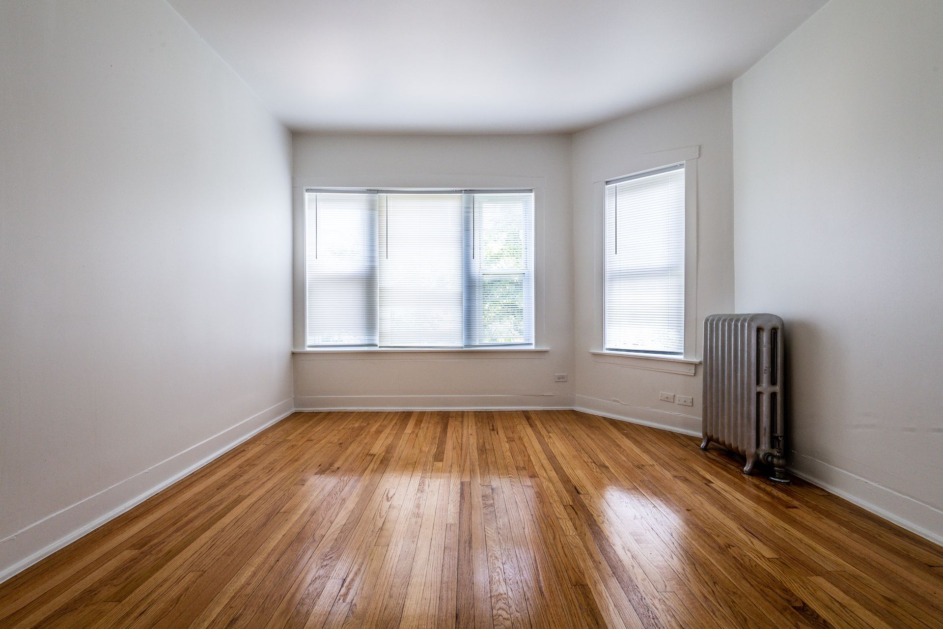 Empty room with hardwood floors, white walls, and two windows with blinds; a radiator is in the corner.
