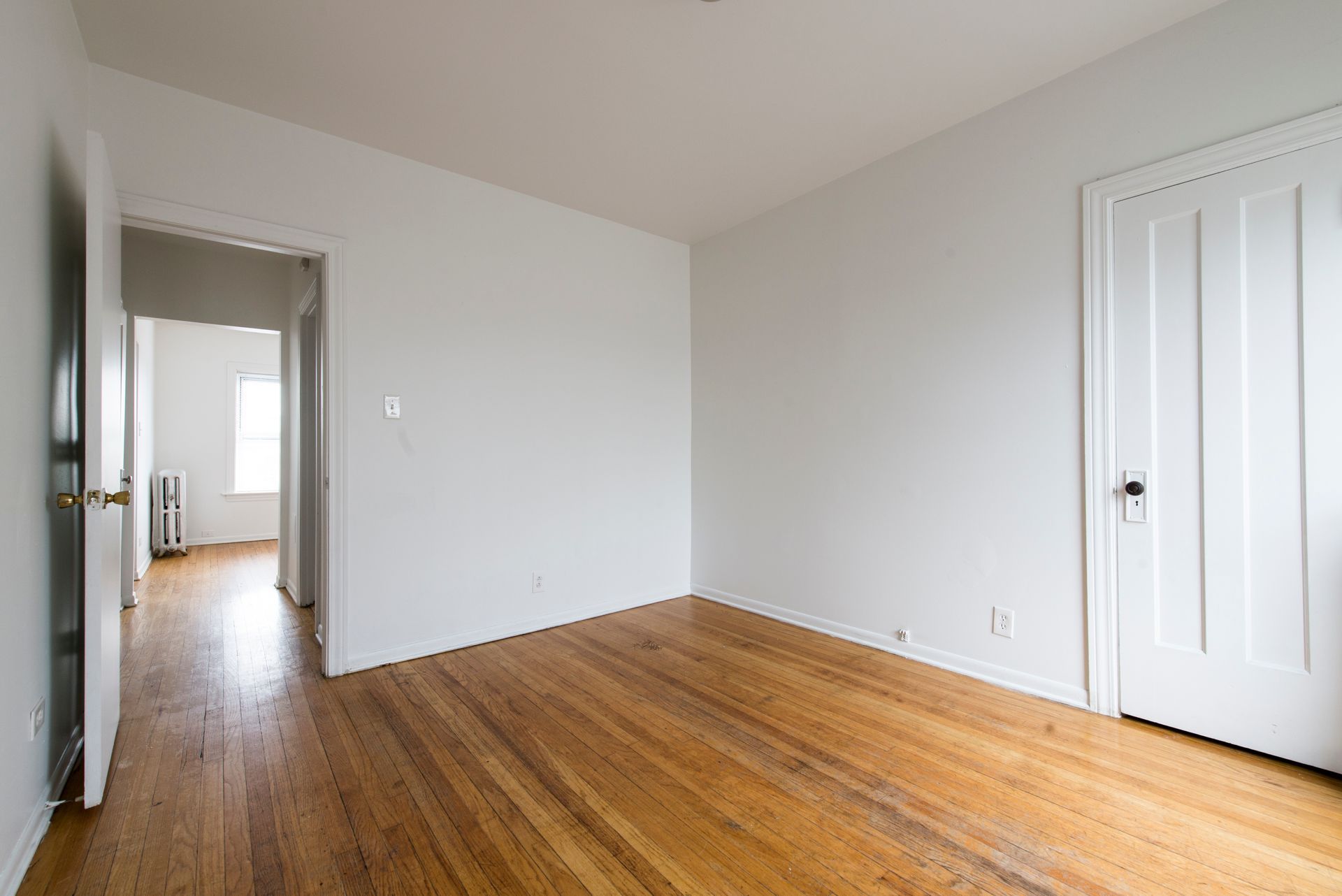 Empty room with wood floors and white walls; doorway to another room on the left, closed white door on the right.