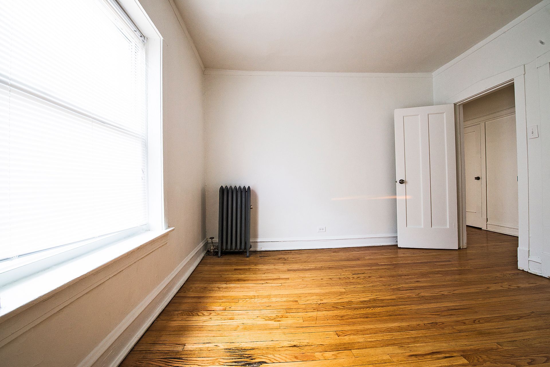 Empty room with hardwood floors, a radiator, and an open doorway; window with blinds.