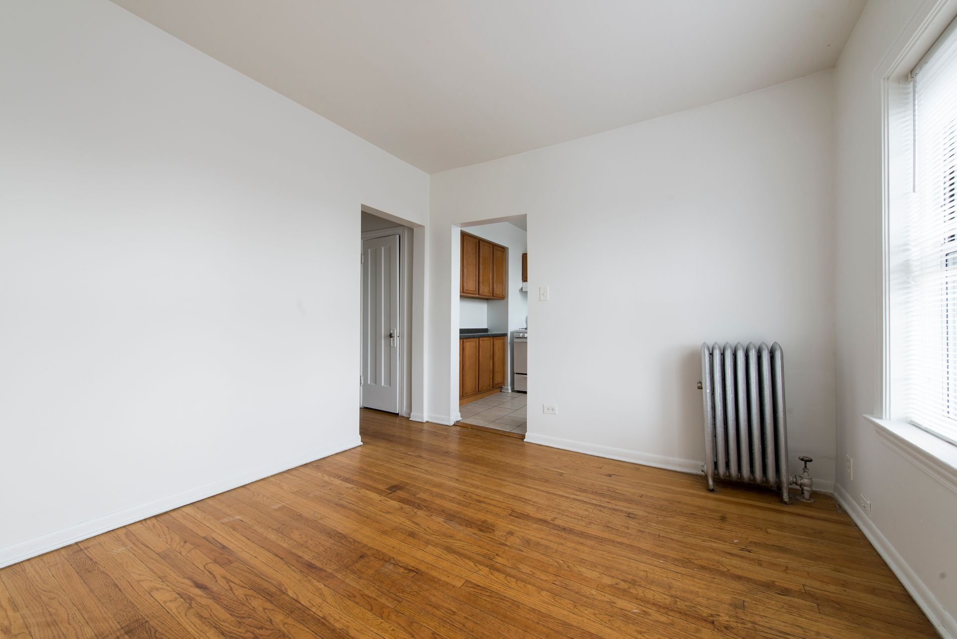 Empty room with hardwood floors, white walls, and a radiator. Doorway leads to a kitchen.