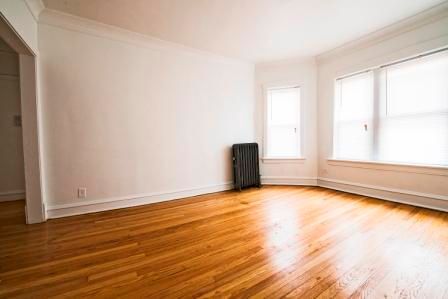 Empty room with hardwood floors, white walls, and a radiator. Two windows provide natural light.