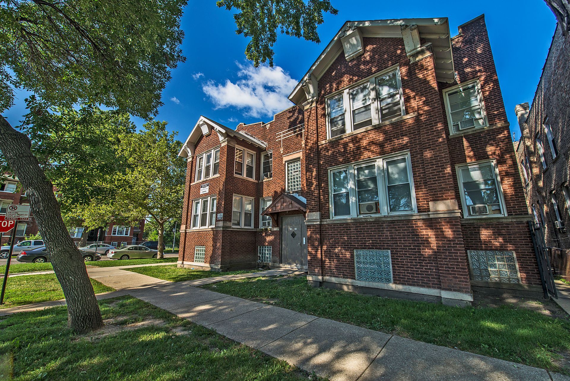 Brick building with multiple windows, surrounded by trees and a sidewalk, under a blue sky.