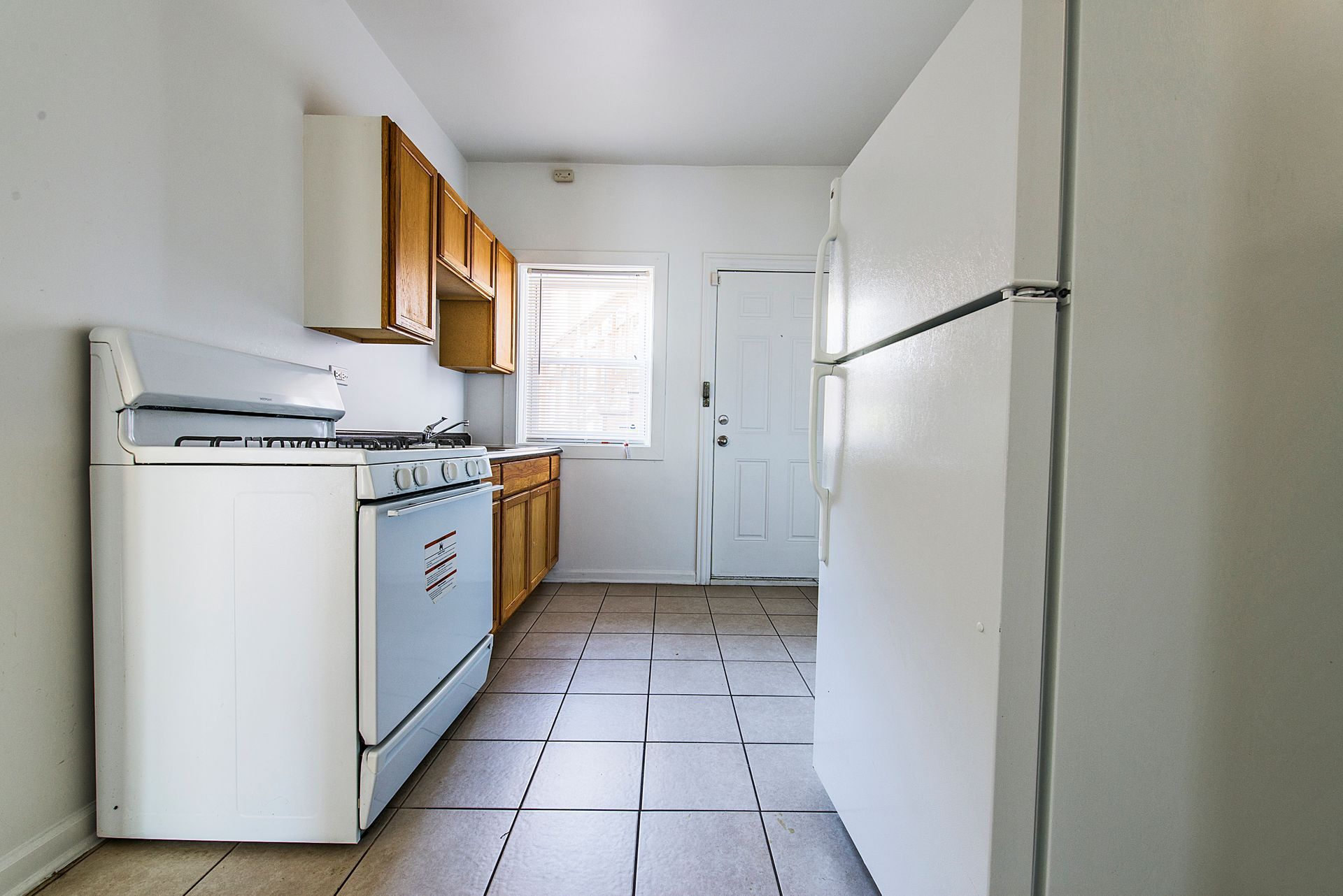 Kitchen with white appliances, wooden cabinets, and tile floor. A window and a door are visible.