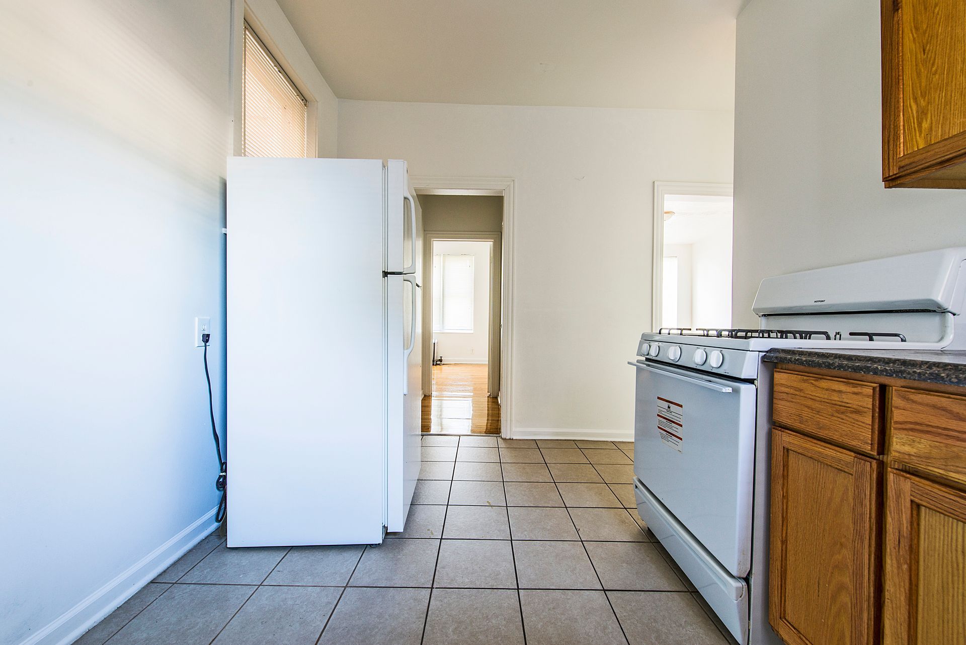 Kitchen with white refrigerator, stove, wooden cabinets, and tiled floor, leading to a hallway.