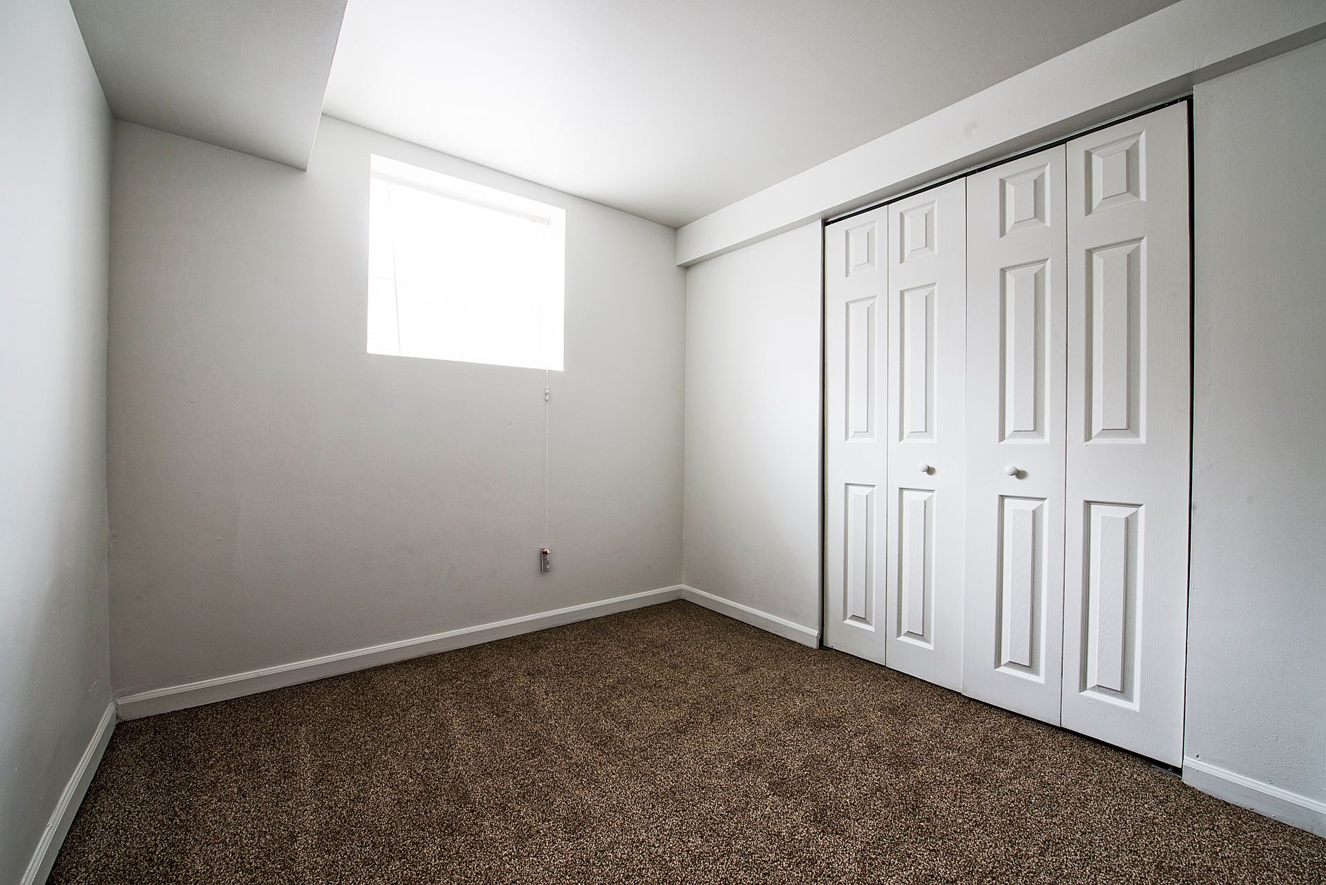 Empty room with white walls, closet, small window, and brown carpet.
