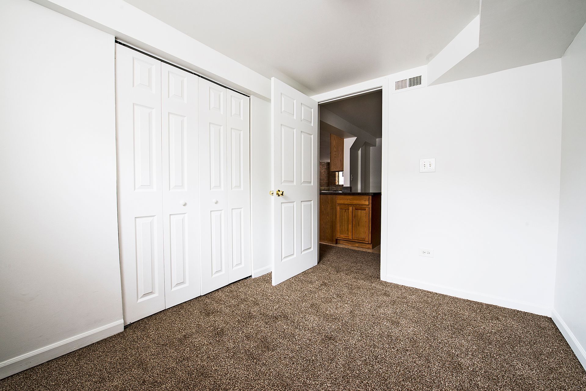 Empty room with brown speckled carpet, white walls, closet, and a doorway to a kitchen.