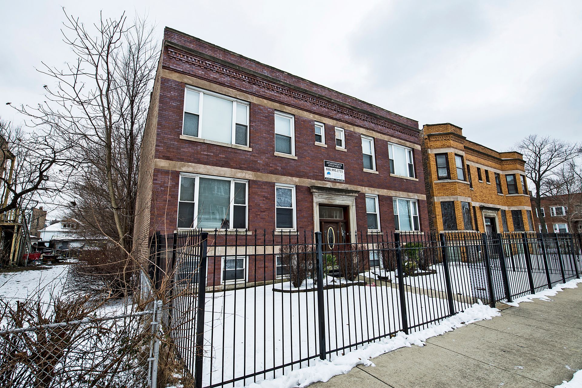 Two-story brick building with black fence, snow-covered ground, and a cloudy sky.