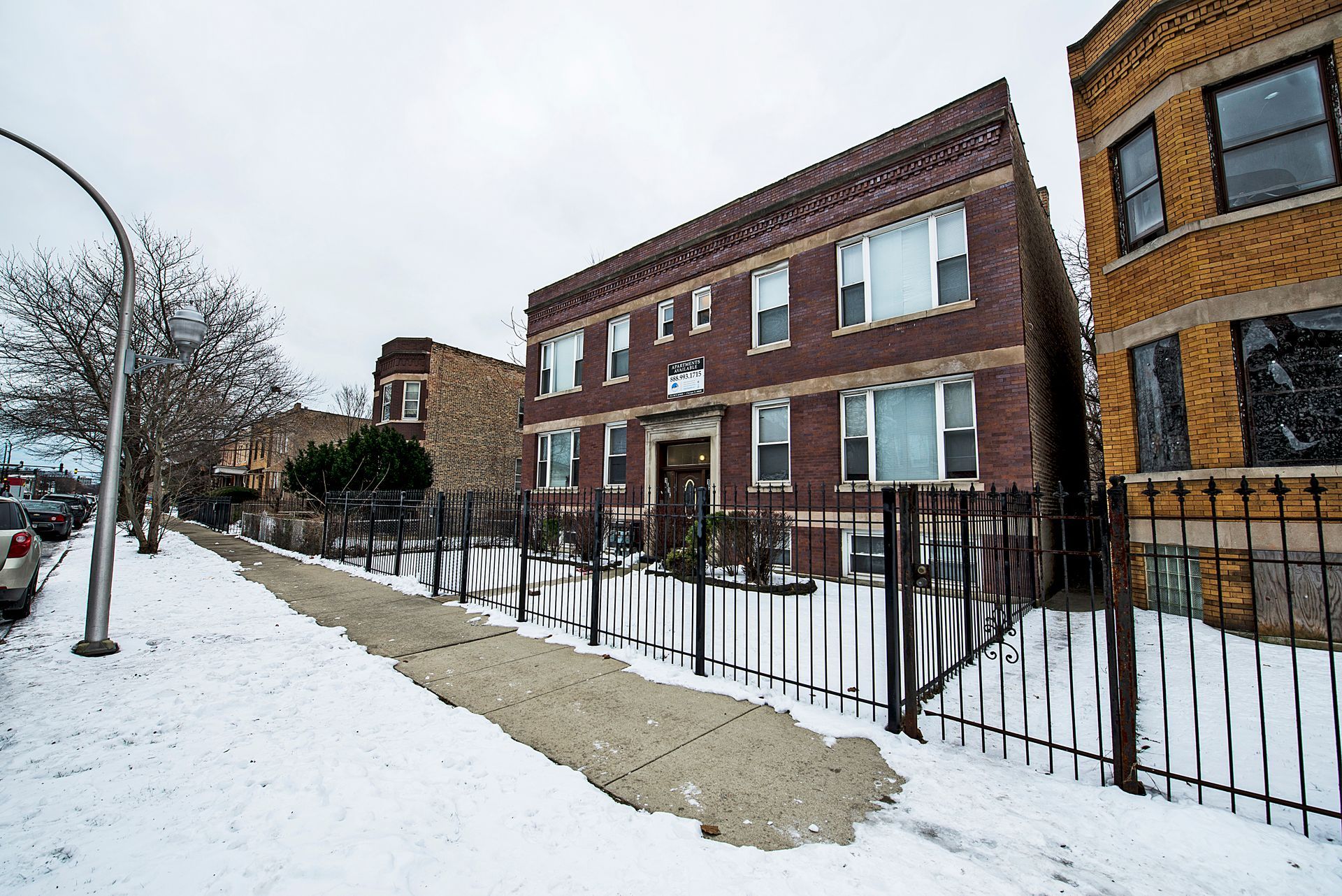 Brick apartment building with snow-covered ground, black fence, and cloudy sky.