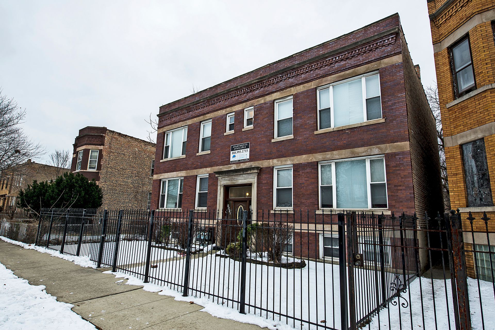 Brick apartment building on a snowy street, with a black fence in front.