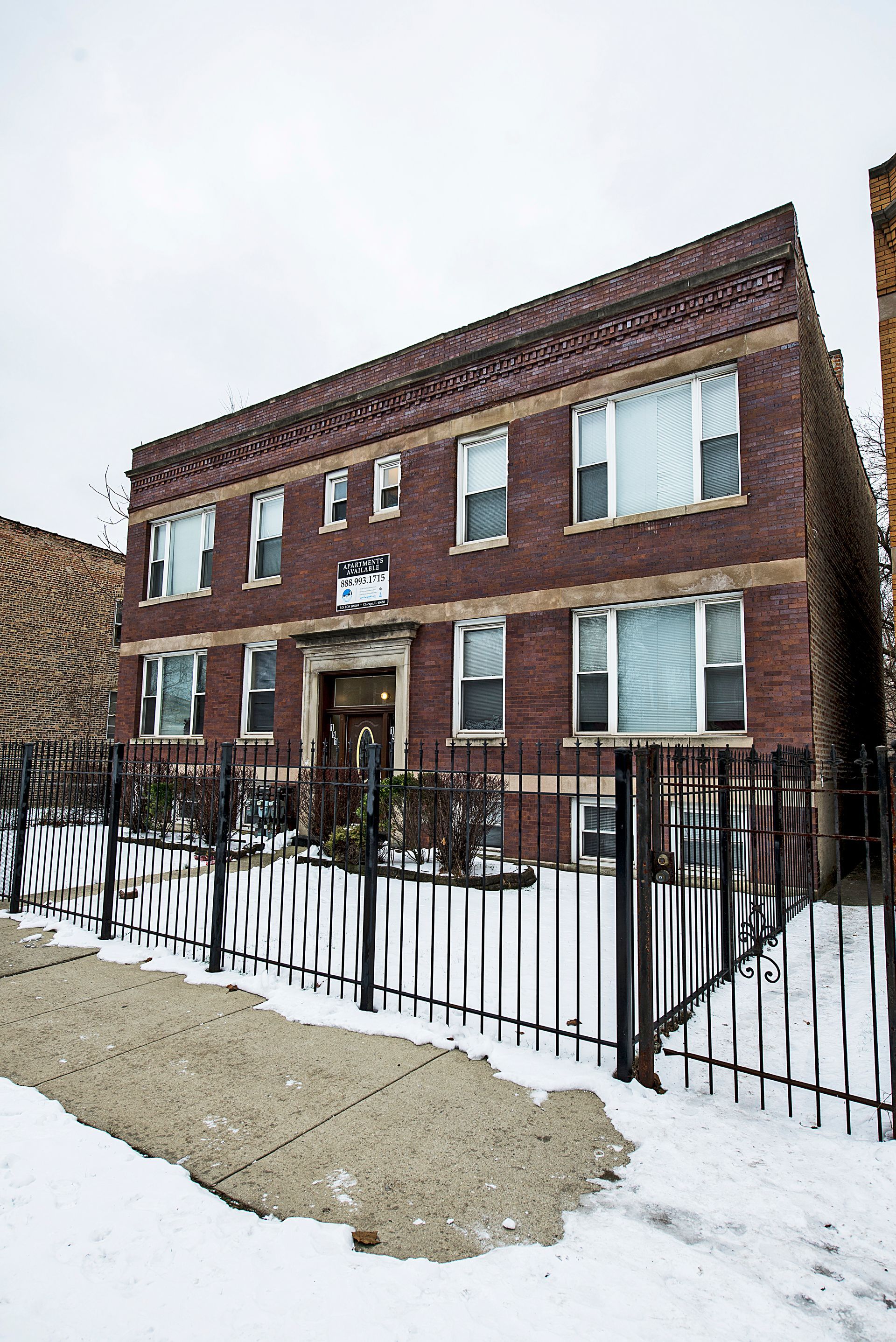 Brick apartment building with snow-covered ground, black fence, overcast sky.