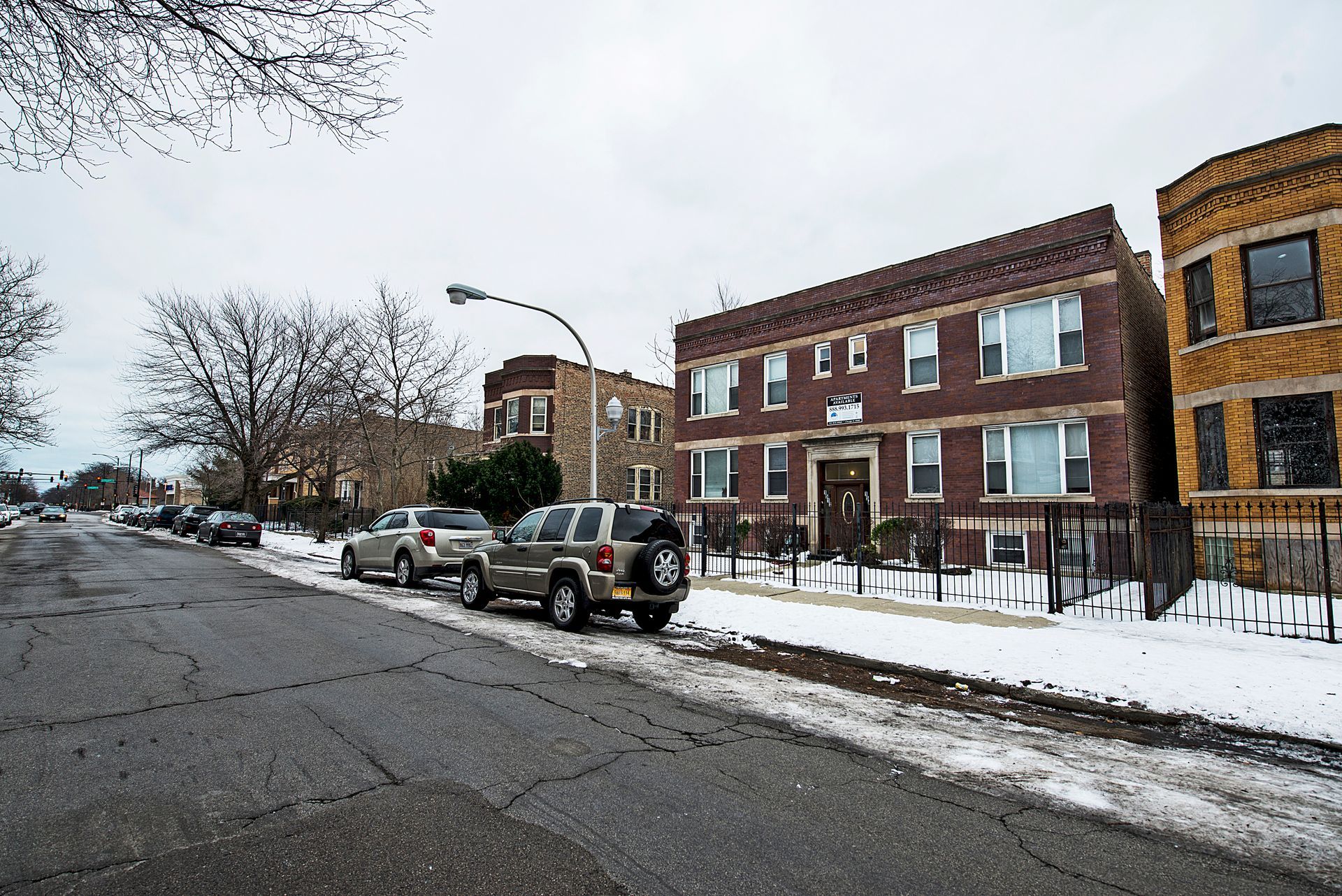 Street scene with brick apartment buildings, parked cars, and snow-covered ground on a cloudy day.