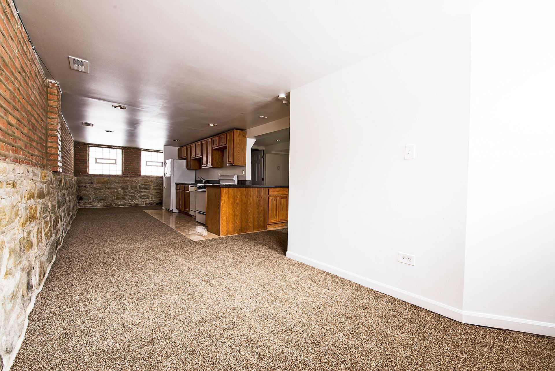 Spacious open-concept living area with exposed brick and stone walls, carpet, and a kitchen in the background.