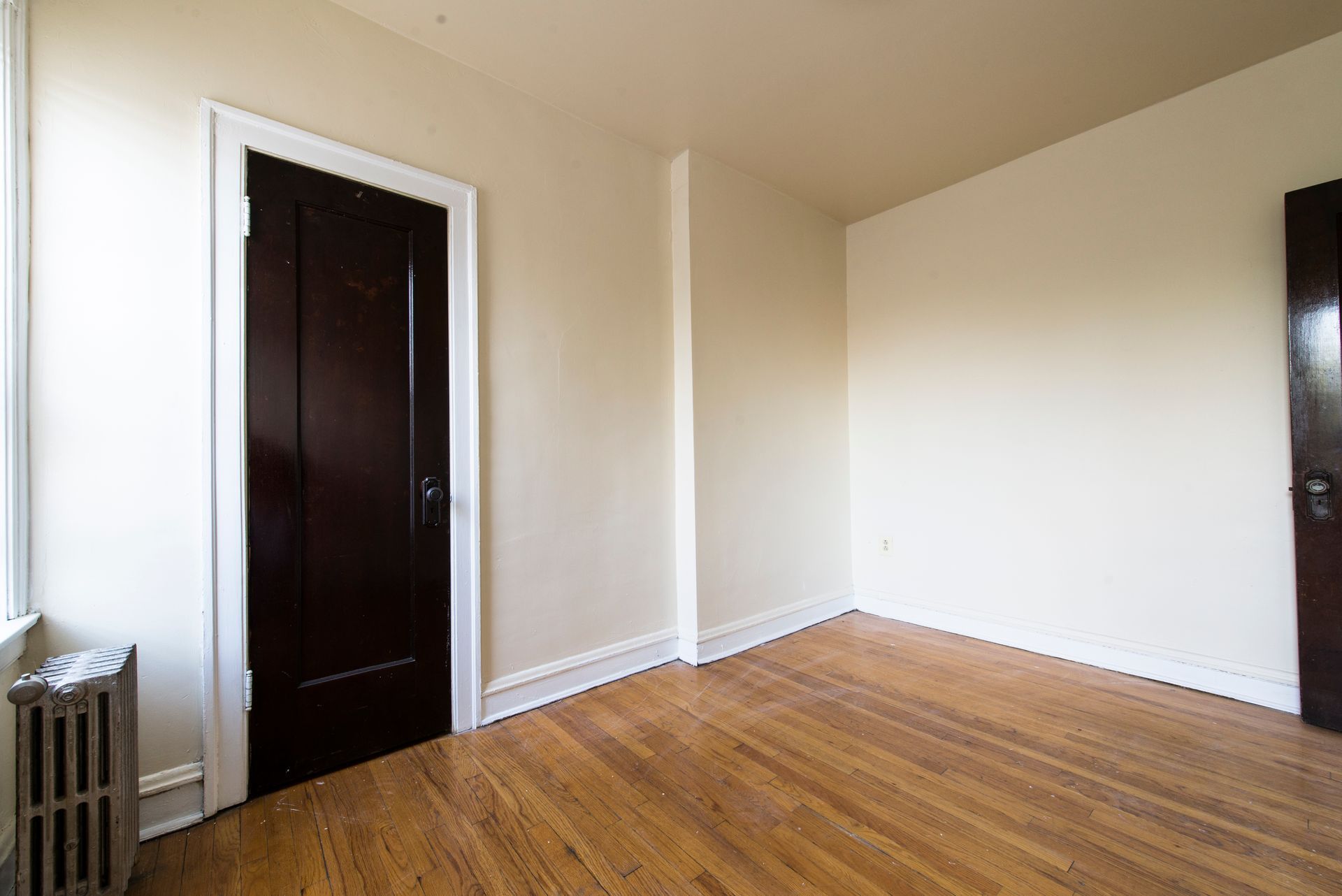 Empty room with hardwood floors, two dark doors, and beige walls.
