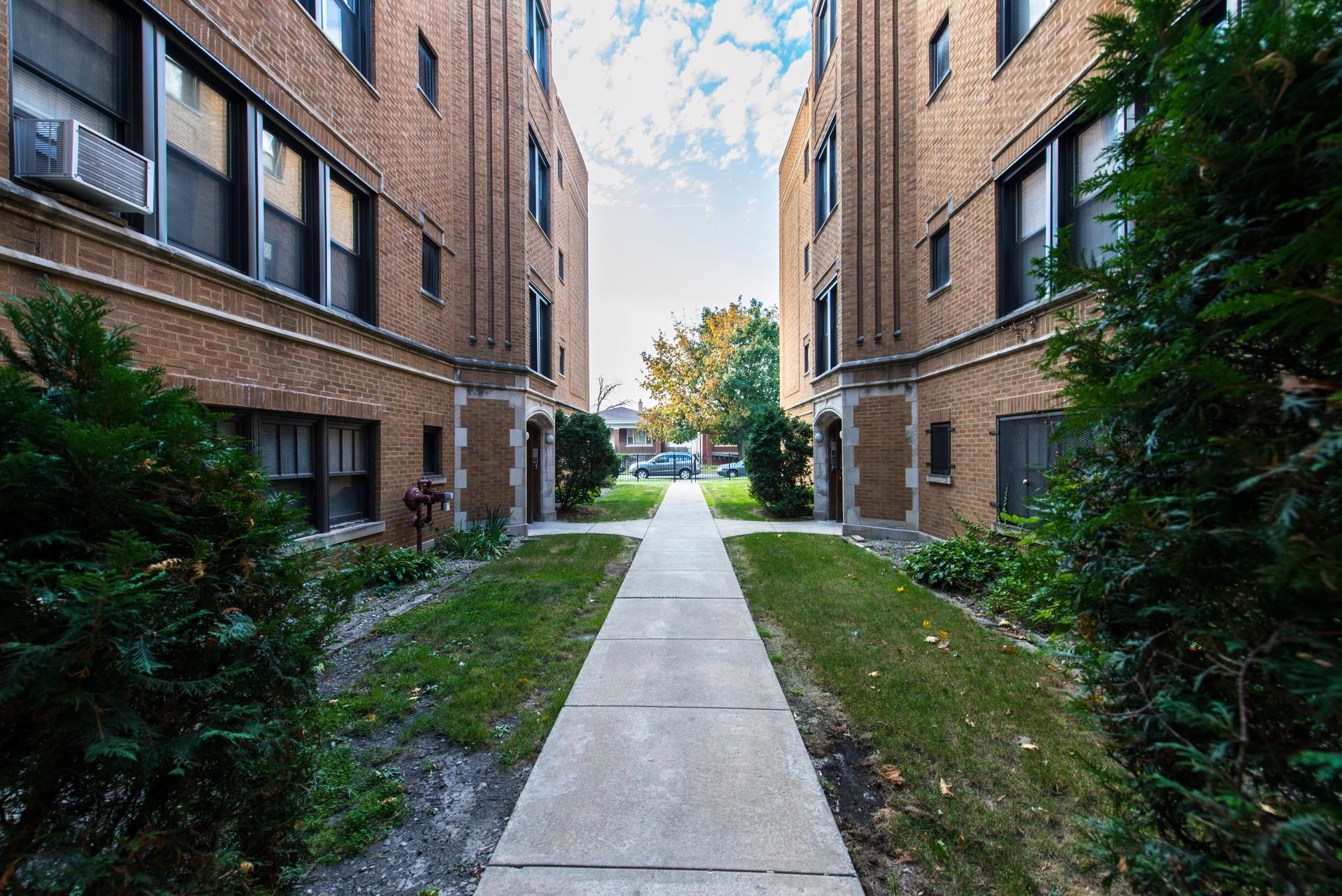 Narrow walkway between two brick apartment buildings. Green grass and shrubbery line the path.