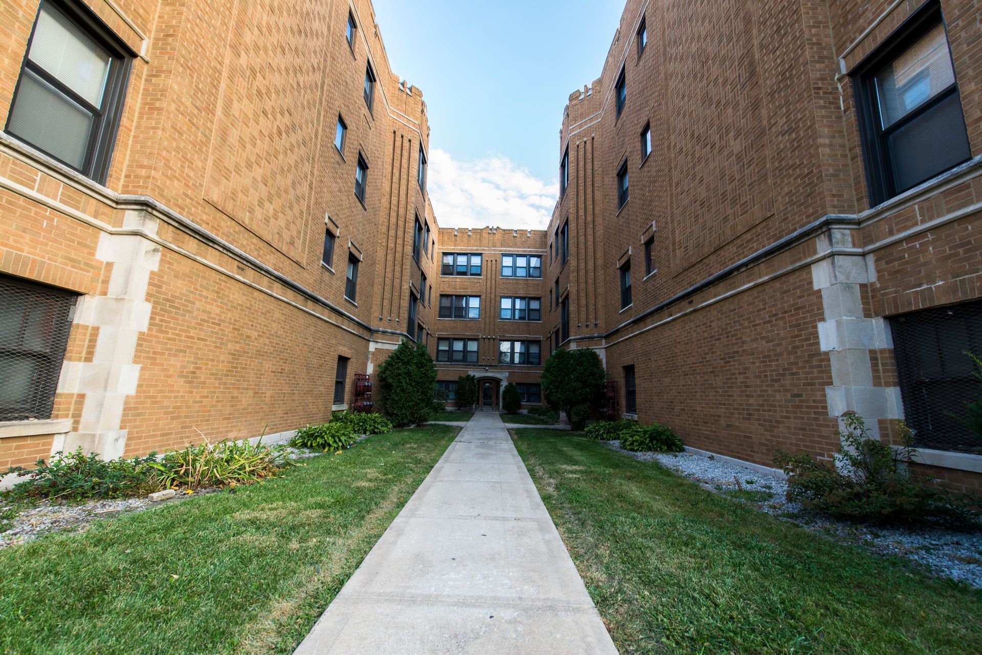 Brick apartment buildings flank a concrete walkway with green grass.