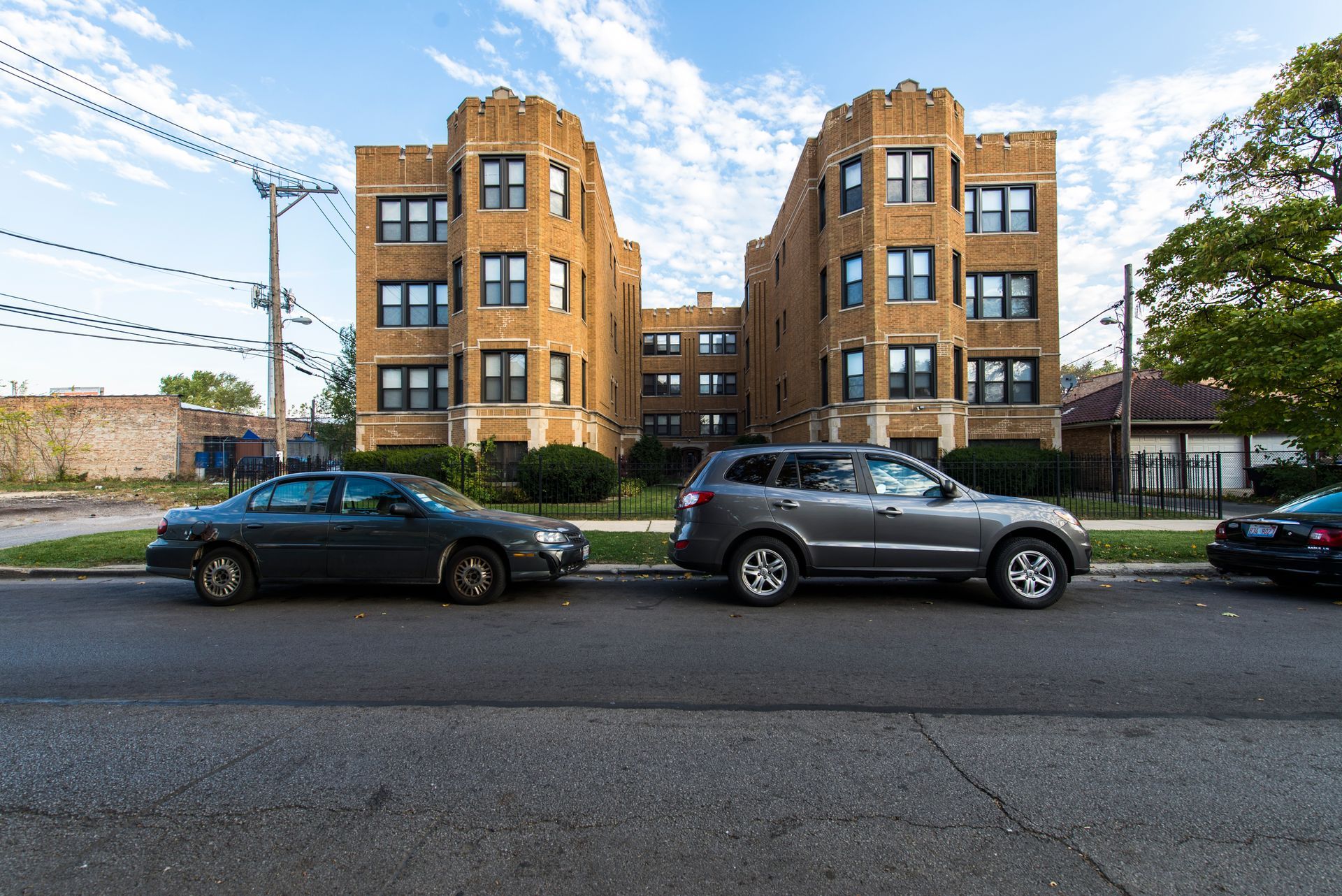 Apartment building with cars parked in front on a city street under a partly cloudy sky.