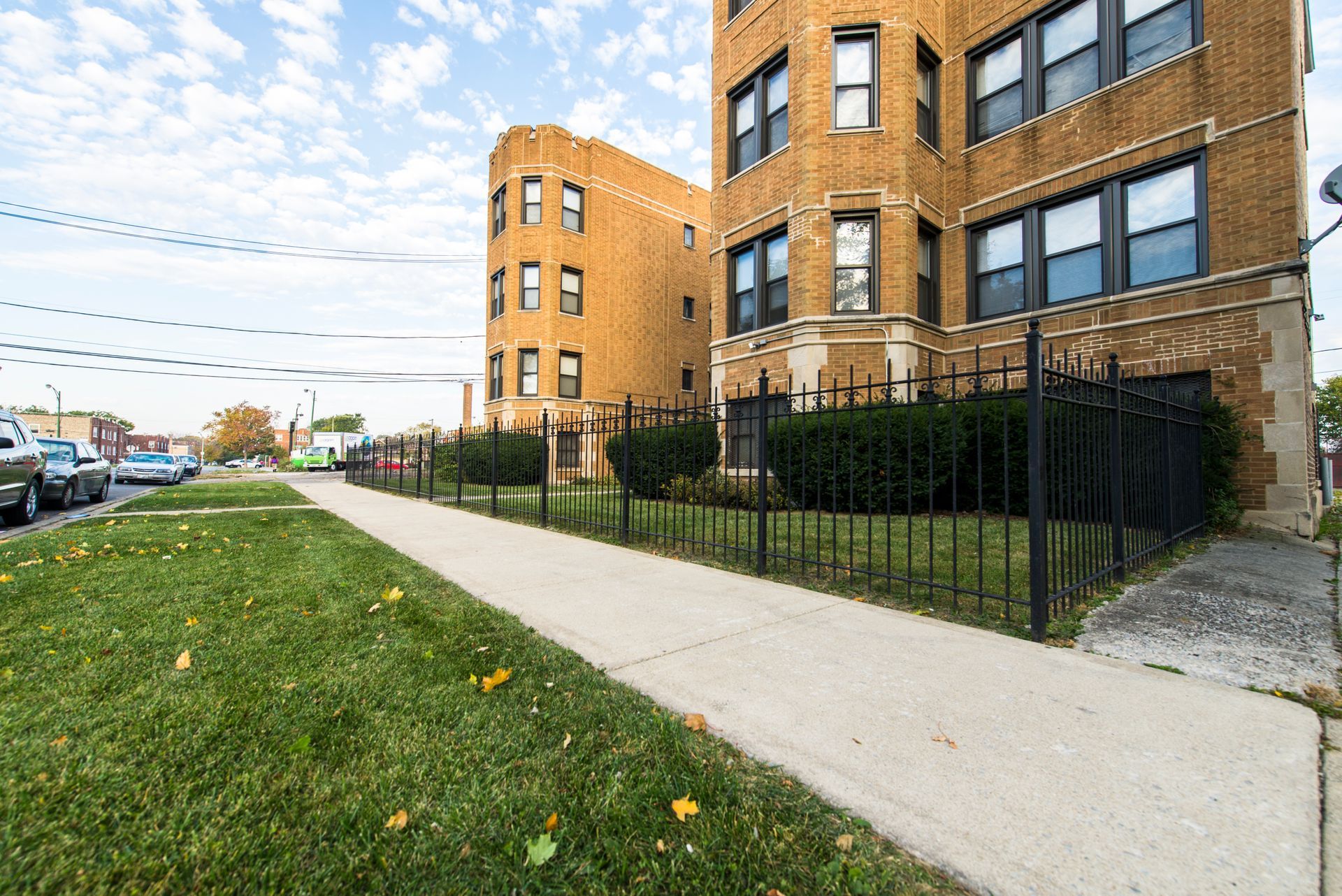 Apartment buildings with a sidewalk, grass, black fence, and cars parked along the street.