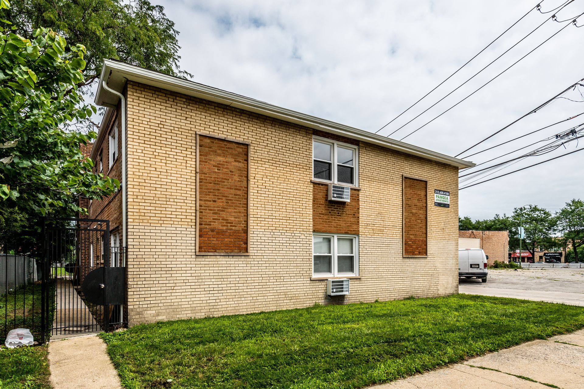Two-story brick building, boarded windows, grass lawn, utility poles, overcast sky.