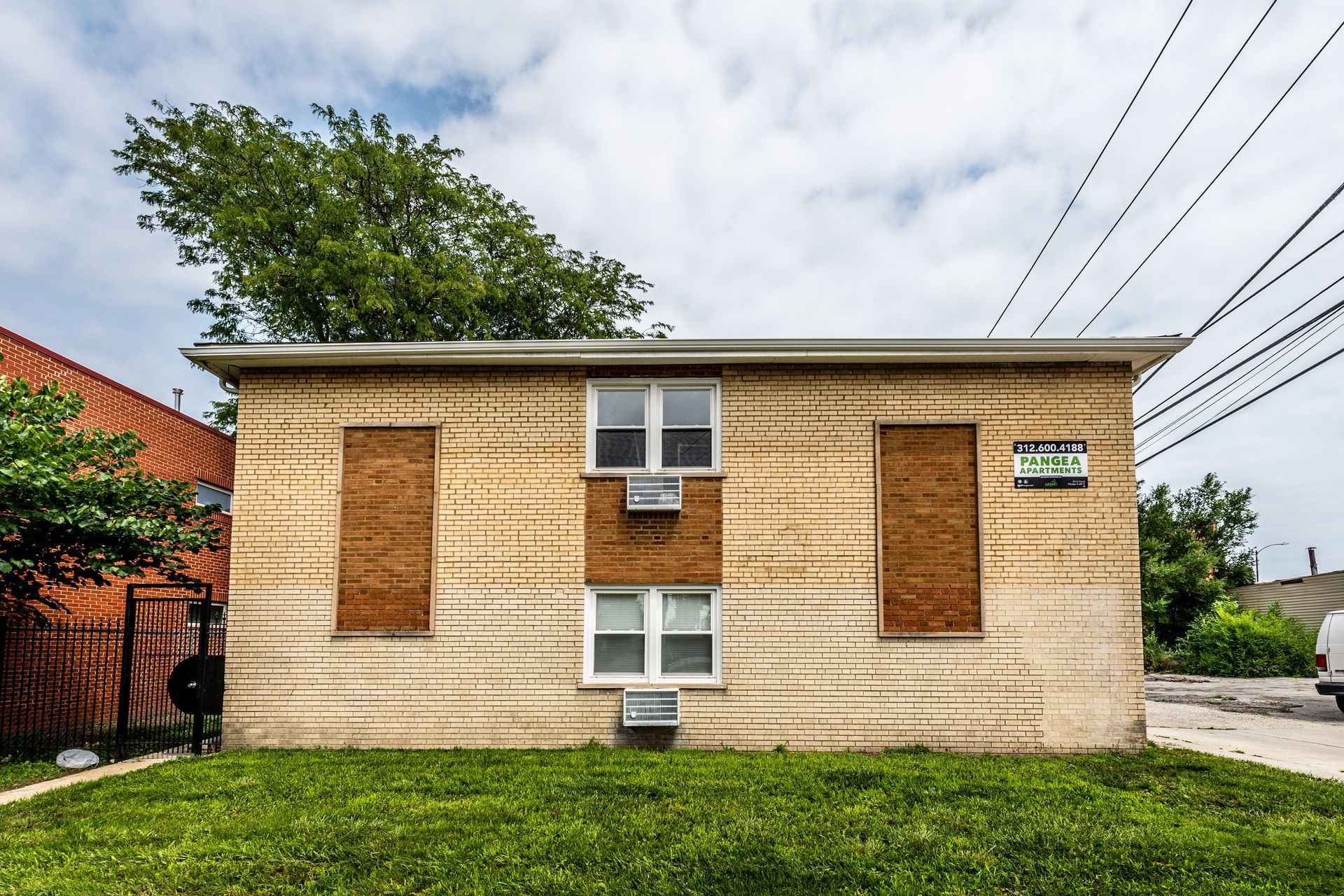 Boarded-up, single-story brick building with two windows and air conditioning units under a cloudy sky.
