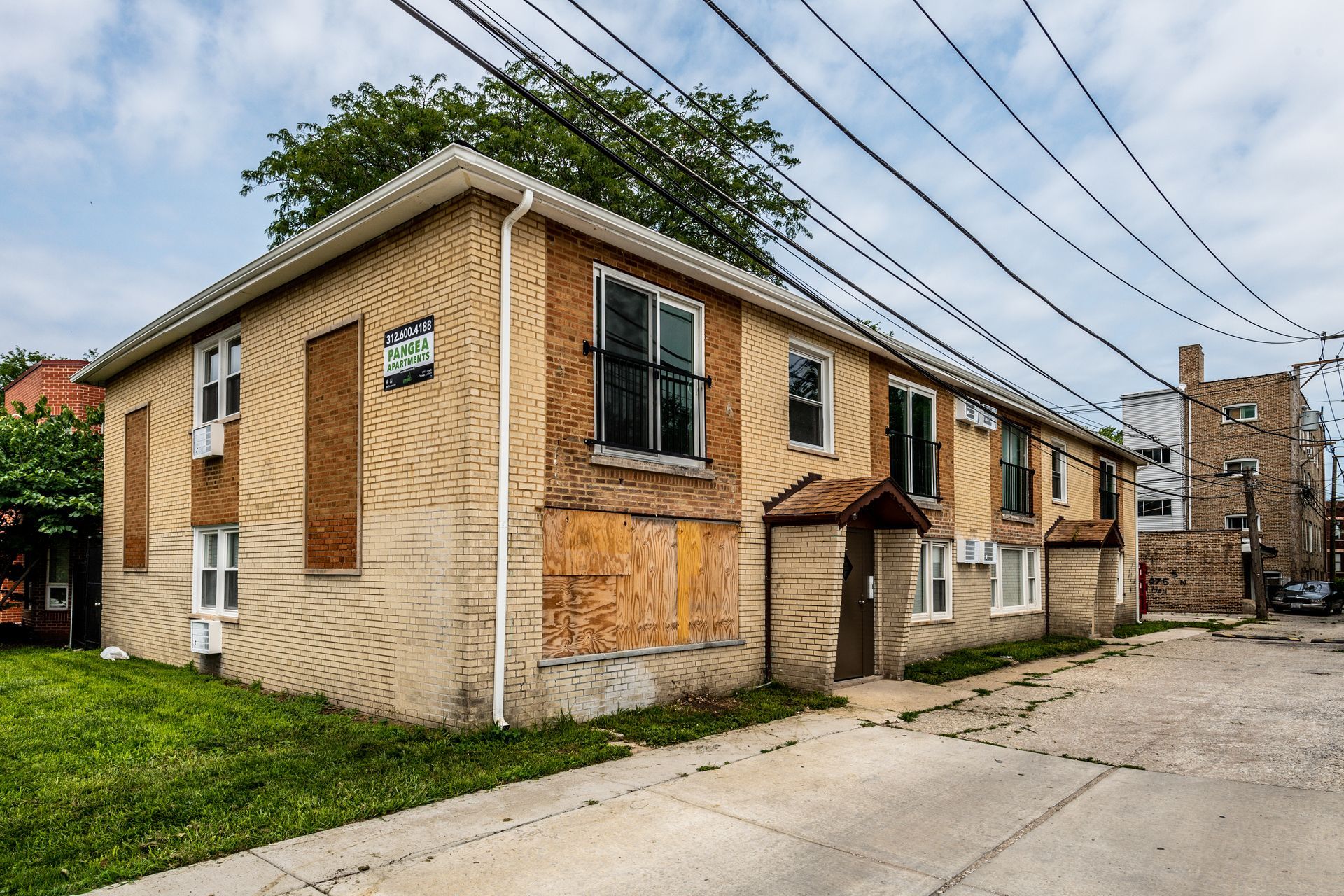 Two-story brick building with boarded windows, surrounded by a cracked sidewalk and power lines.