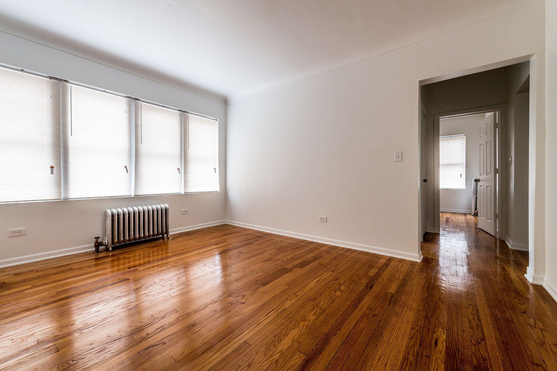Empty room with hardwood floors, windows with blinds, and doorway to a hallway.