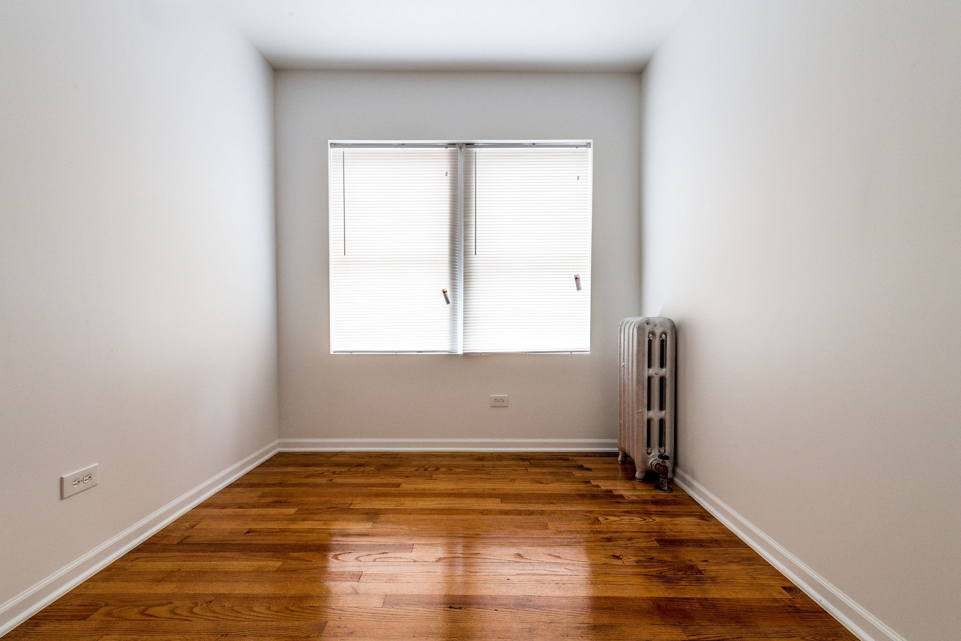 Empty room with wooden floor, window with blinds, and radiator.