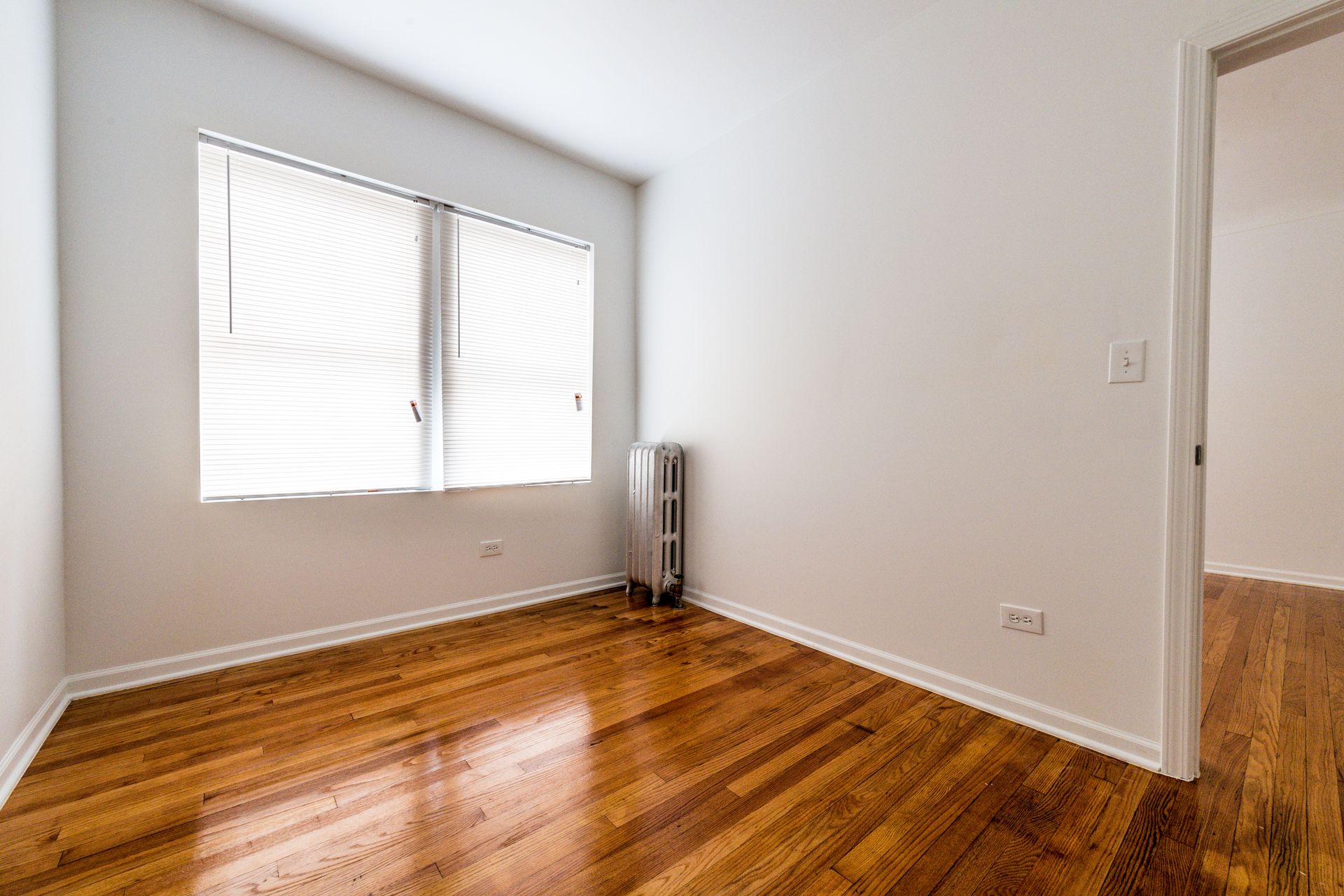 Empty room with hardwood floors, a window with blinds, and a doorway.