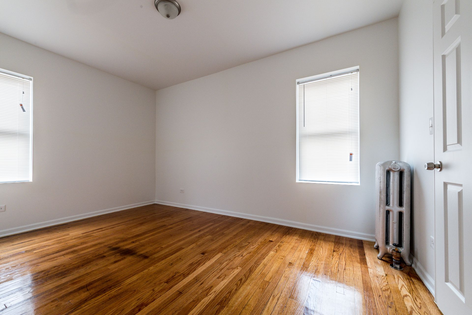 Empty room with hardwood floors, two windows with blinds, and a radiator.
