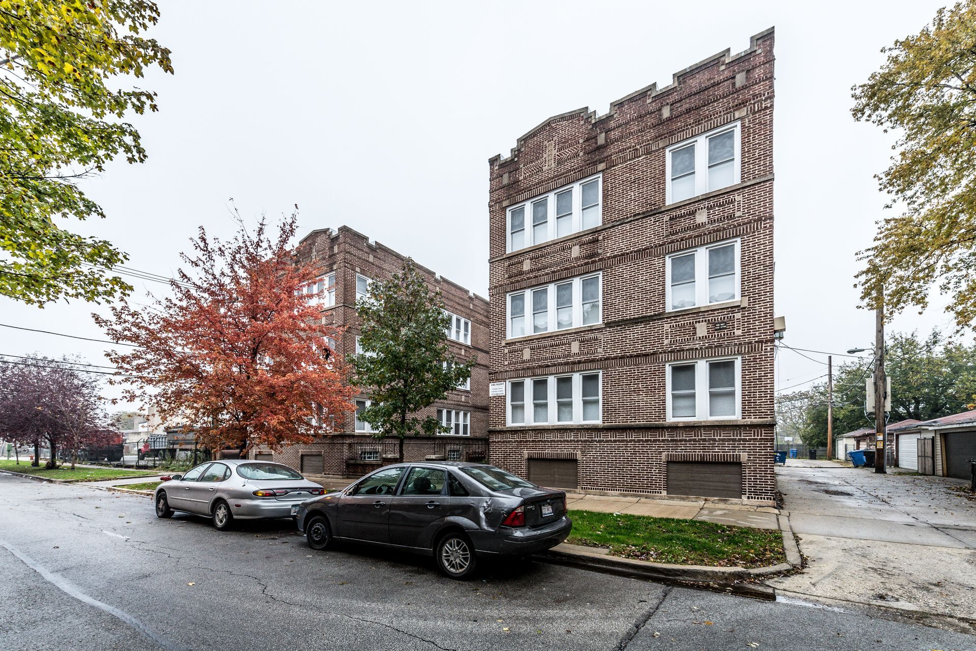Brick apartment building with parked cars on a city street; trees with autumn leaves.