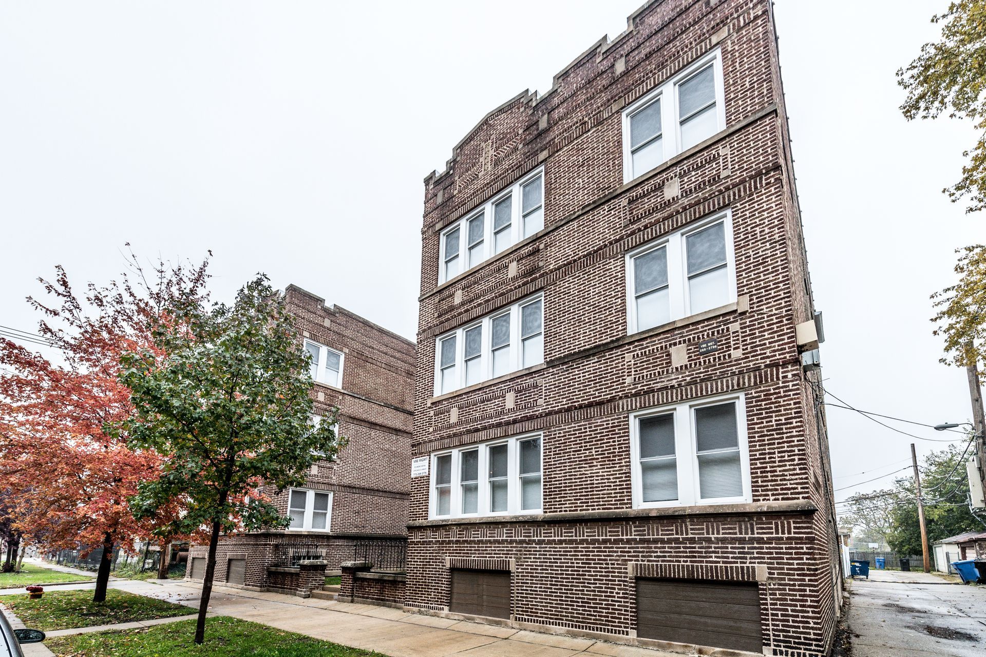 Brick apartment building with white-framed windows on a cloudy day. Trees with red and green leaves frame the building.