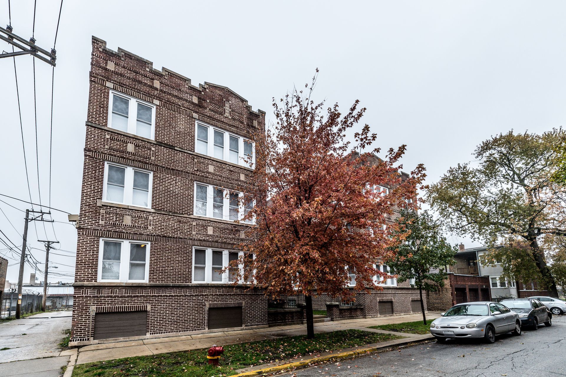 Brick apartment building on a street with parked cars and a tree with red leaves. Overcast sky.