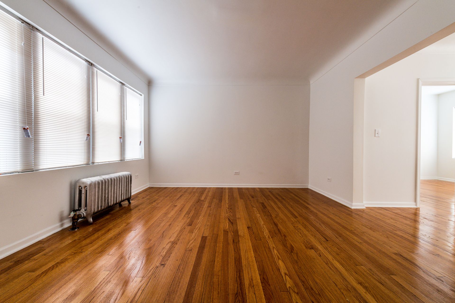 Empty room with hardwood floor, radiator, and large window with blinds.