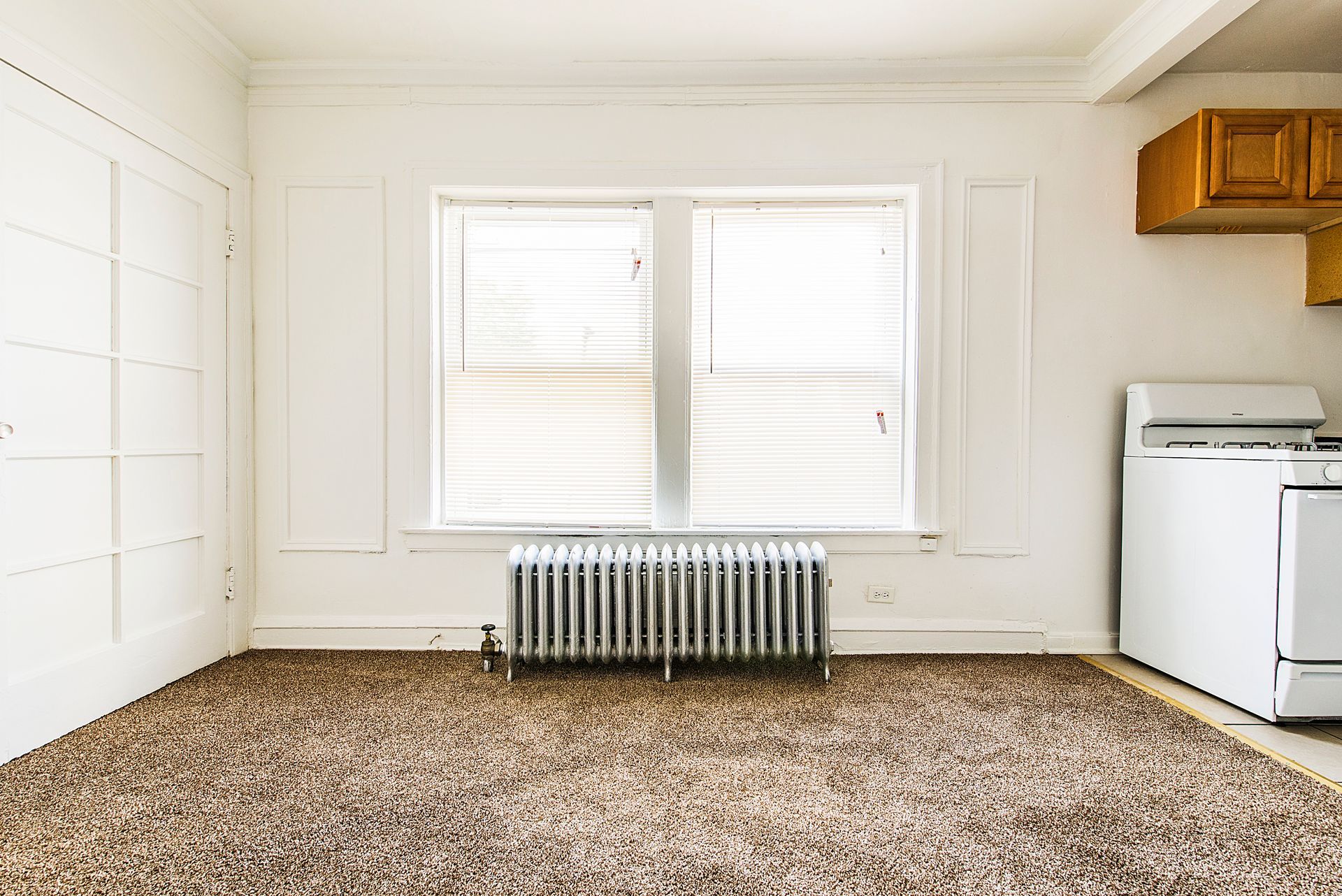 Empty room with a window, radiator, and a stove. Tan carpet and white walls.