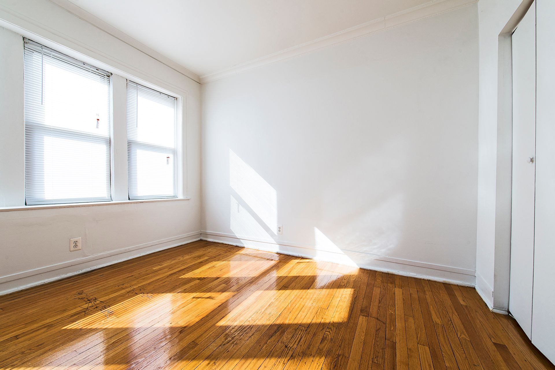 Empty living room with wood floors, brick fireplace, and large windows. Sunlight streams in.