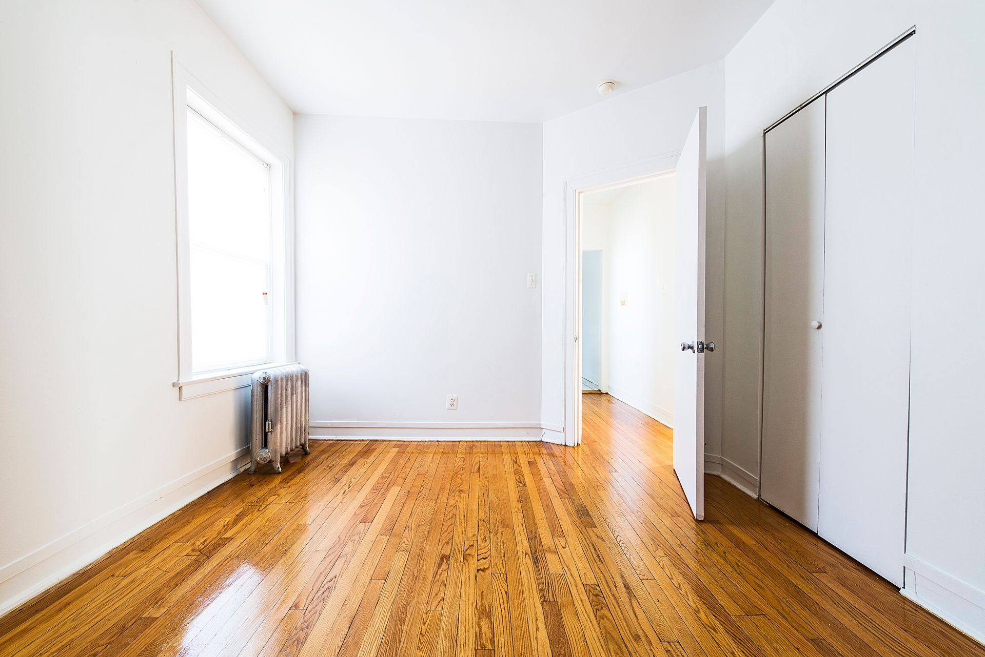 Empty living room with wood floors, brick fireplace, and large windows. Sunlight streams in.