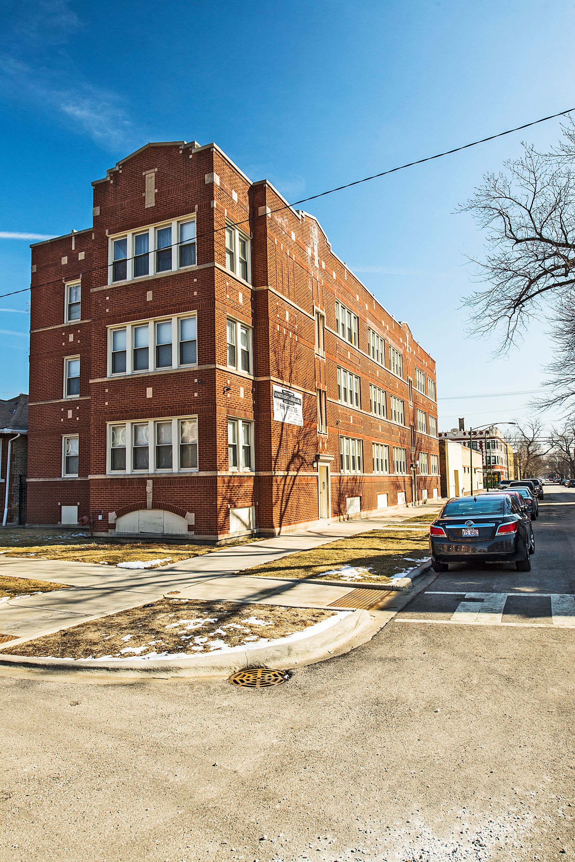 Red brick apartment building on a street corner, blue sky overhead. A car parked nearby.