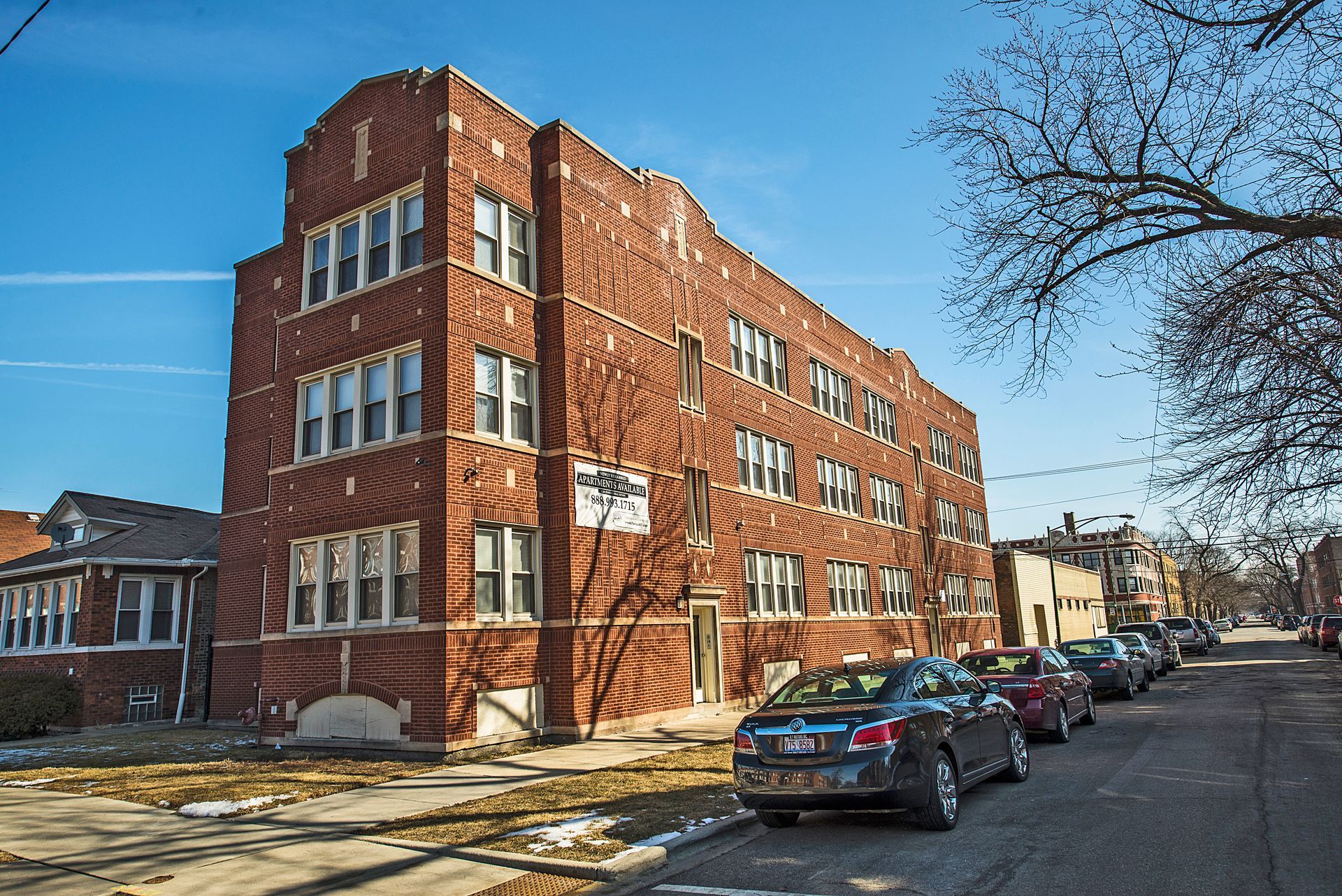 Three-story brick apartment building on a residential street; cars parked along curb.