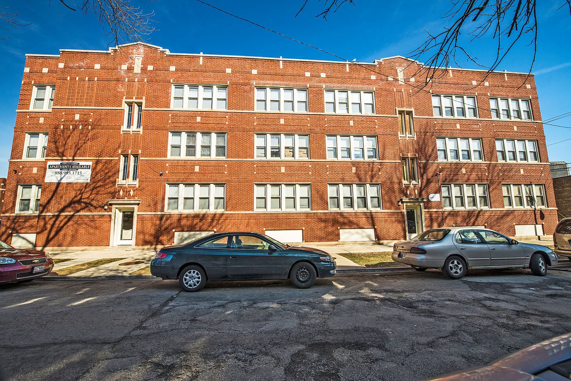 Red brick apartment building with parked cars on a sunny street.