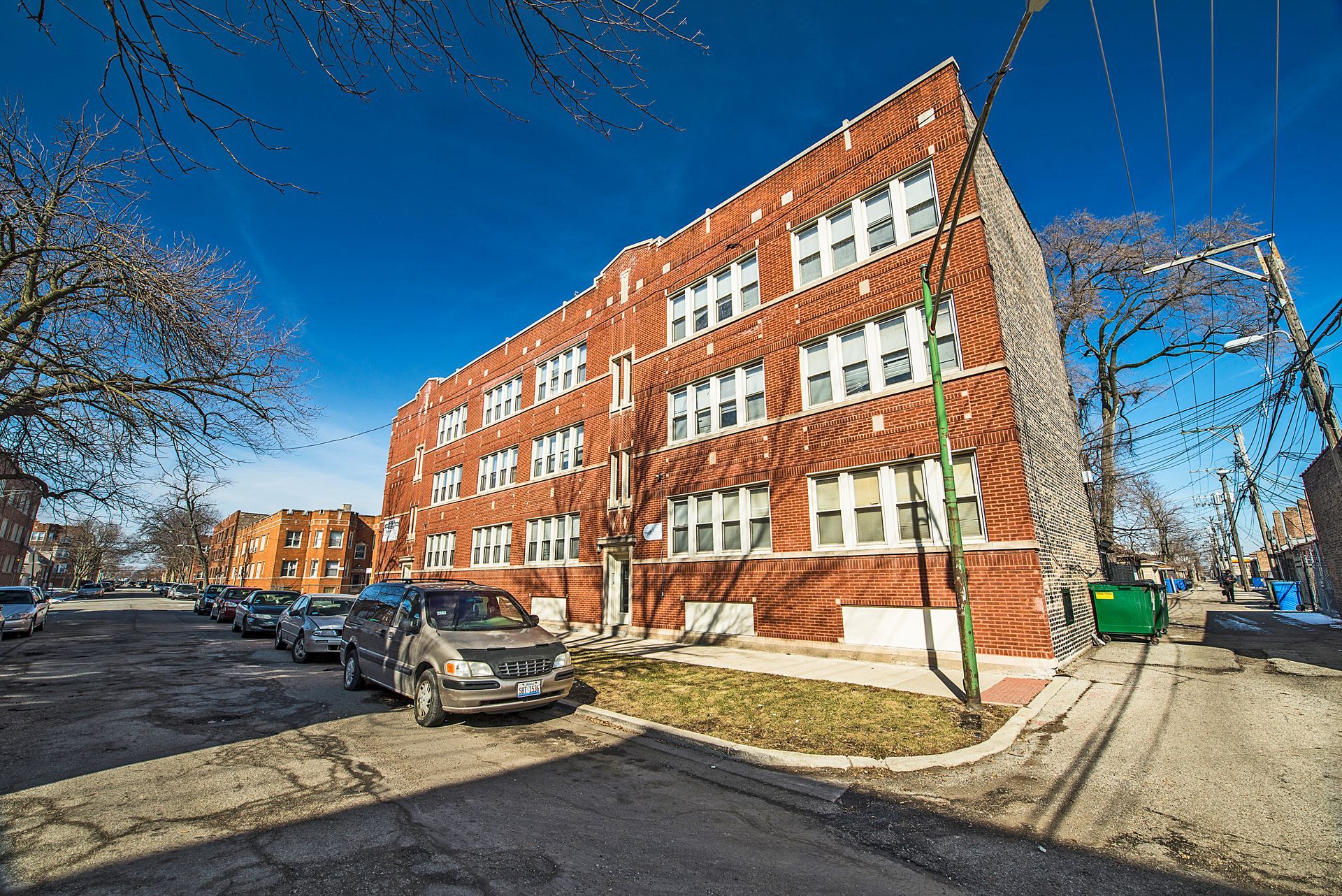 Brick apartment building on a street with parked cars and power lines against a clear blue sky.