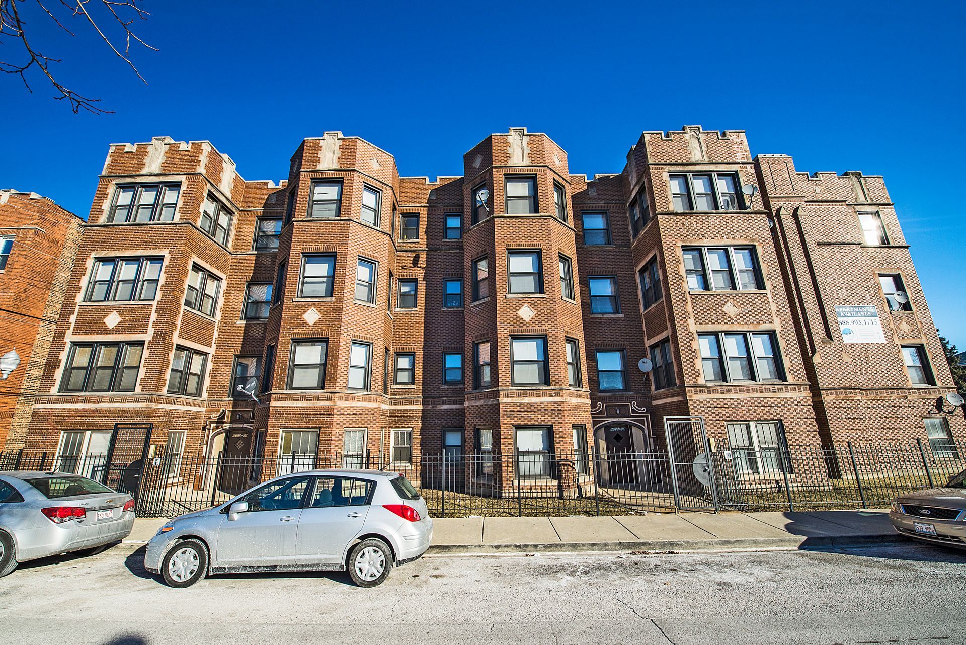 Multi-story brick apartment building with cars parked in front under a clear blue sky.