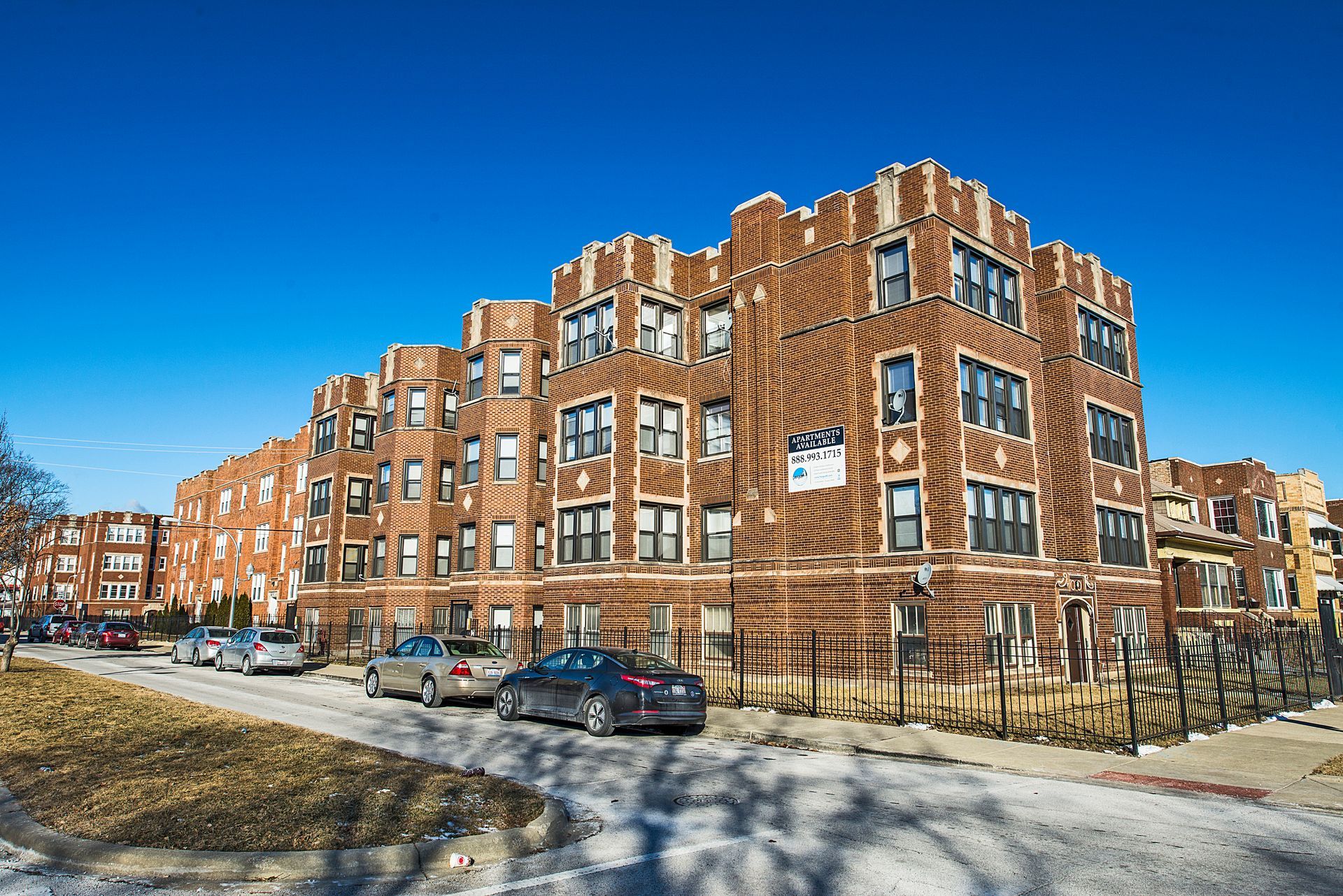 Brick apartment building under a bright blue sky; cars parked on street in front.